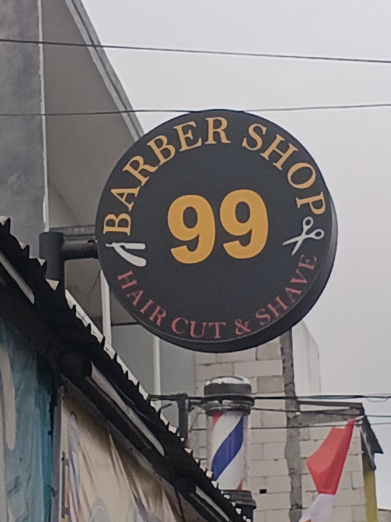 A circular barber shop sign with the text "BARBER SHOP 99 HAIR CUT & SHAVE" is mounted on a building in the city of Surabaya, Indonesia. The sign is dark gray with gold lettering, except for the "99" which is in yellow, and two white barber tools, a razor and scissors, on either side of the numbers. Below the sign, a traditional barber pole with red, white, and blue stripes is visible, along with a red and white flag. The background shows a gray, overcast sky and a building with a textured facade. The perspective suggests the photo was taken from a slightly upward angle, looking at the sign.