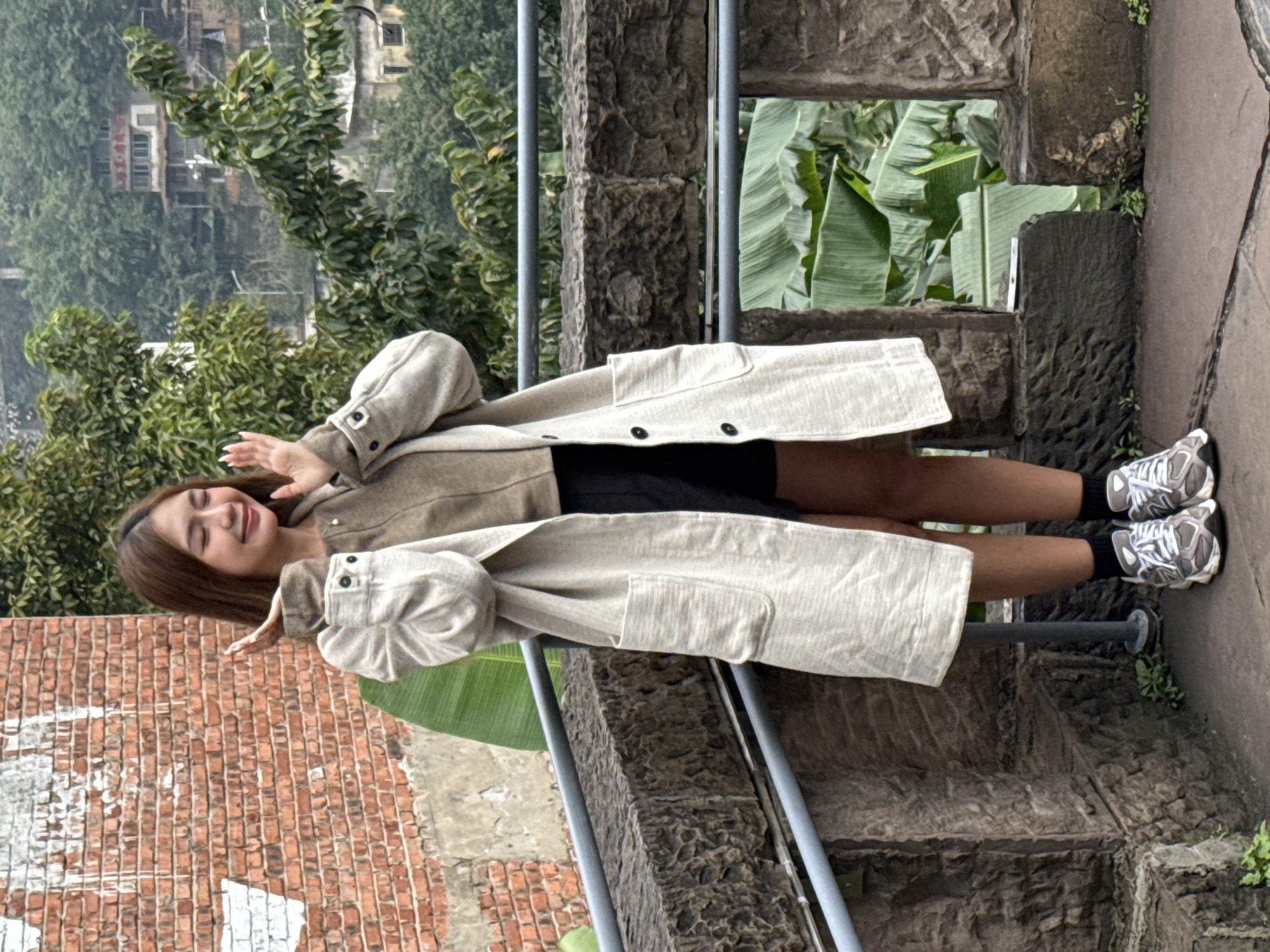 A young adult female stands on a stone-paved outdoor terrace within the Nan'an District, China. She is positioned slightly right of center, facing to her right with eyes closed and a wide smile, her hands raised to chest height with palms forward and fingers splayed. She wears a light brown long-sleeved top, black shorts, a long, light beige textured overcoat with dark buttons and front pockets, black socks, and grey and white athletic shoes. To her immediate left is a weathered red brick wall with irregular white paint marks. A dark, rough-hewn stone parapet, topped with two grey metal rails, forms a barrier across the midground. Below and beyond the parapet, large green banana leaves are prominent. The background features a dense, green, hilly landscape interspersed with various multi-story buildings, one displaying red characters on its facade, indicative of an urban environment.