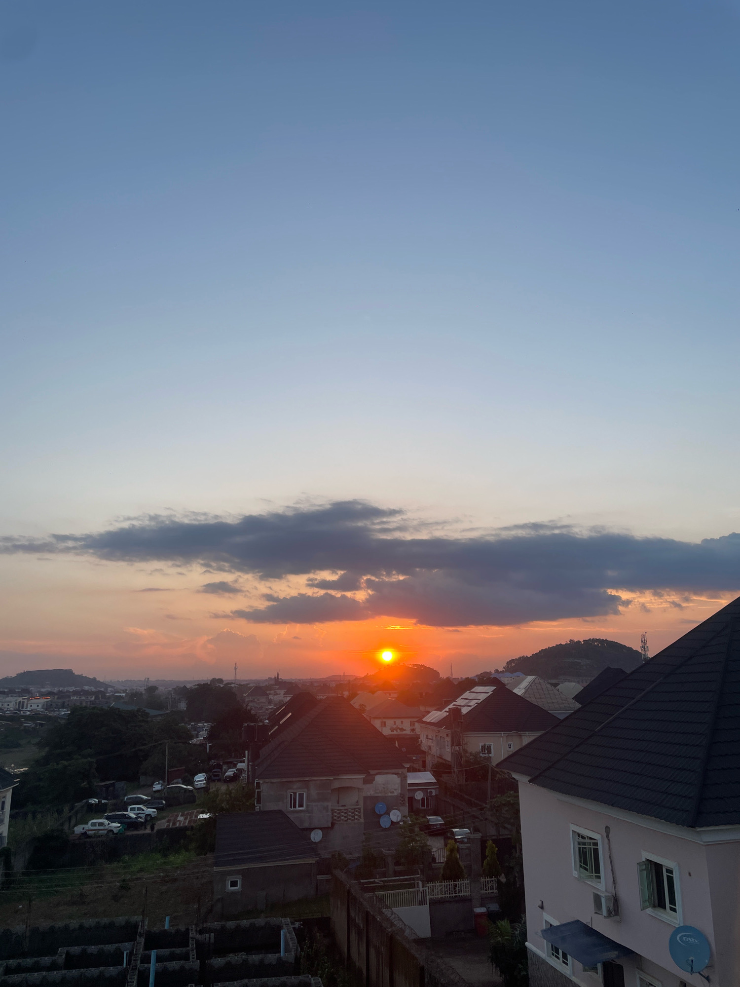 The image captures a sunset over a residential area in Abuja, Nigeria. The sky is a gradient of soft blues at the top, transitioning to warmer hues of orange and pink near the horizon where the sun is setting, partially obscured by clouds. The sun appears as a bright, glowing orb. In the foreground, rooftops of houses are visible, predominantly with dark-colored tiles. Some houses have visible windows and air conditioning units. In the middle ground, more houses and buildings are seen, interspersed with trees and vegetation. A road with several parked cars, including white SUVs and sedans, is also present. The background features rolling hills with some structures and antennas on them, contributing to the cityscape of Abuja. The overall scene depicts a tranquil evening in a suburban or semi-urban environment. There are no people visible in the image.