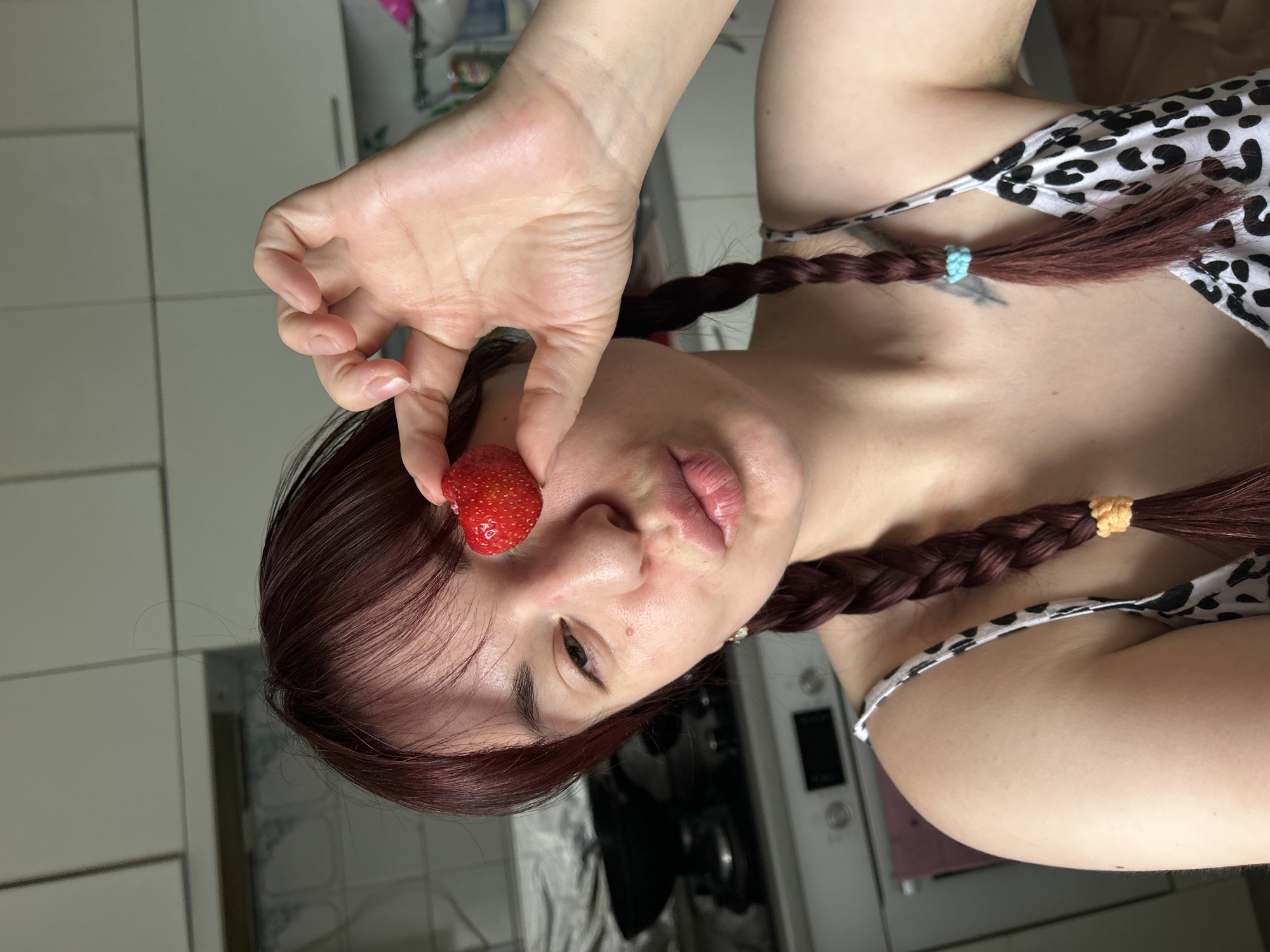 In a medium shot taken from a low angle, a young woman with auburn hair styled in two braids stands in what appears to be a kitchen. She is holding a single strawberry up to her right eye, obscuring it, and puckering her lips. Her gaze is directed at the camera. She is wearing a white top with a black leopard print. The background shows kitchen cabinets and appliances, suggesting an indoor domestic setting. The lighting appears to be natural, coming from an unseen window. The overall impression is of a playful and informal moment captured on camera. The location is not specified.