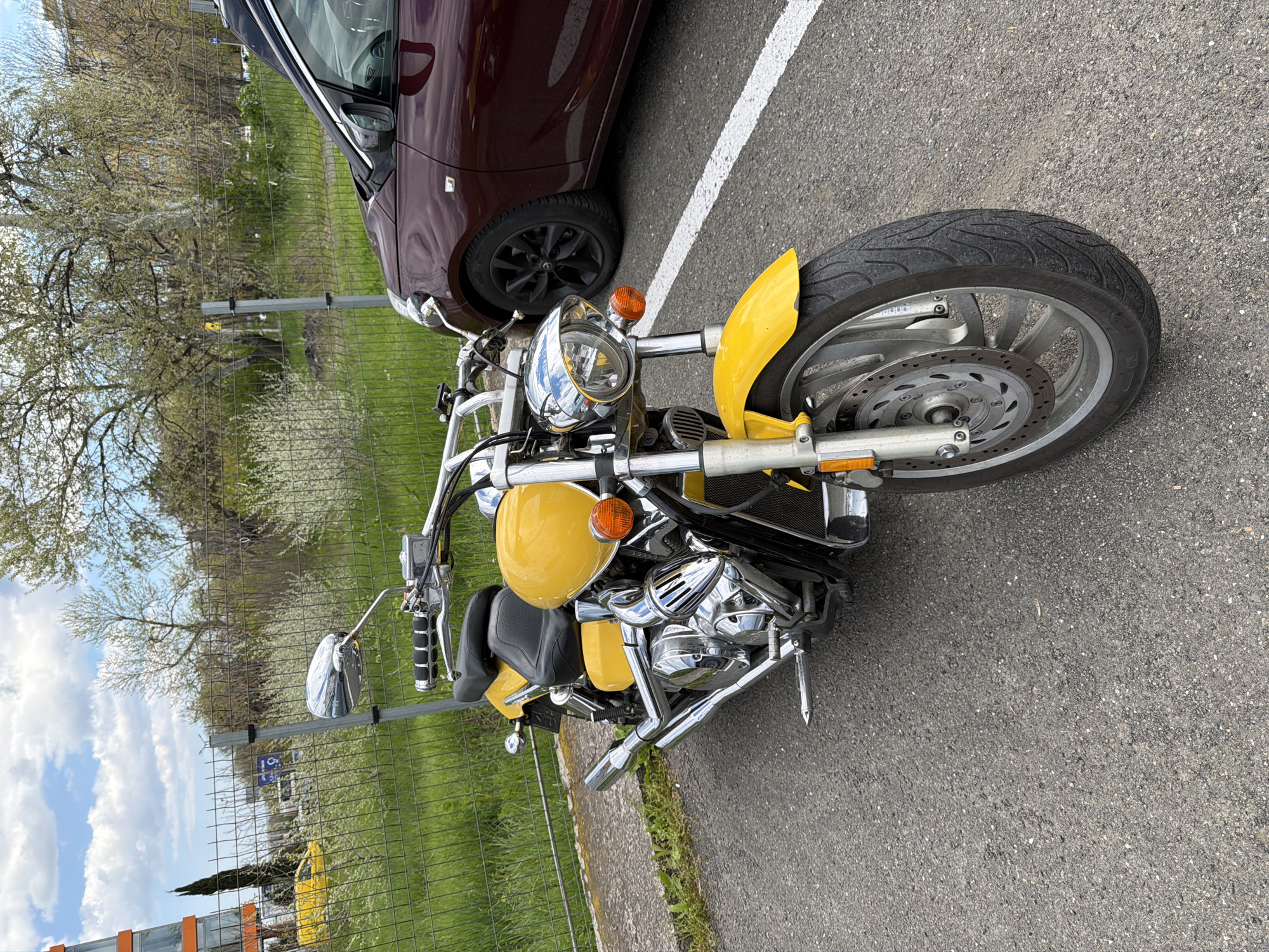 The image, originally oriented horizontally and rotated 90 degrees counter-clockwise, depicts a yellow motorcycle and a dark red car parked on an asphalt surface under a bright sky.
In the foreground and mid-ground, a striking yellow and chrome cruiser-style motorcycle is prominently featured. It boasts a large, shiny chrome headlight, chrome handlebars with reflective mirrors, and a detailed chrome engine and exhaust system. The front fender is yellow, matching the main body, and it has black dual seats. A large front wheel with a disc brake system is clearly visible. The tire sidewall shows the brand "DUNLOP" and model "D401".
To the left of the motorcycle, a dark red or maroon car is partially seen, showing its front wheel, fender, and a portion of its side mirror and hood. A white parking line is painted on the asphalt between the vehicles.
The background features a wire or chain-link fence separating the parking area from a natural landscape. Beyond the fence, there is a vibrant green grassy field, dotted with trees displaying light green spring foliage and some white blossoms, suggesting it's springtime. Above the tree line, a blue sky with scattered white clouds is visible. Notable details in the background include a small yellow post and a blue road sign with a white 'P' in a circle, indicating a parking area, along with what appears to be an arrow and another symbol below it.
The scene is illuminated by bright daylight, suggesting a sunny or partly cloudy day. No people are present, and the vehicles appear to be stationary and parked.