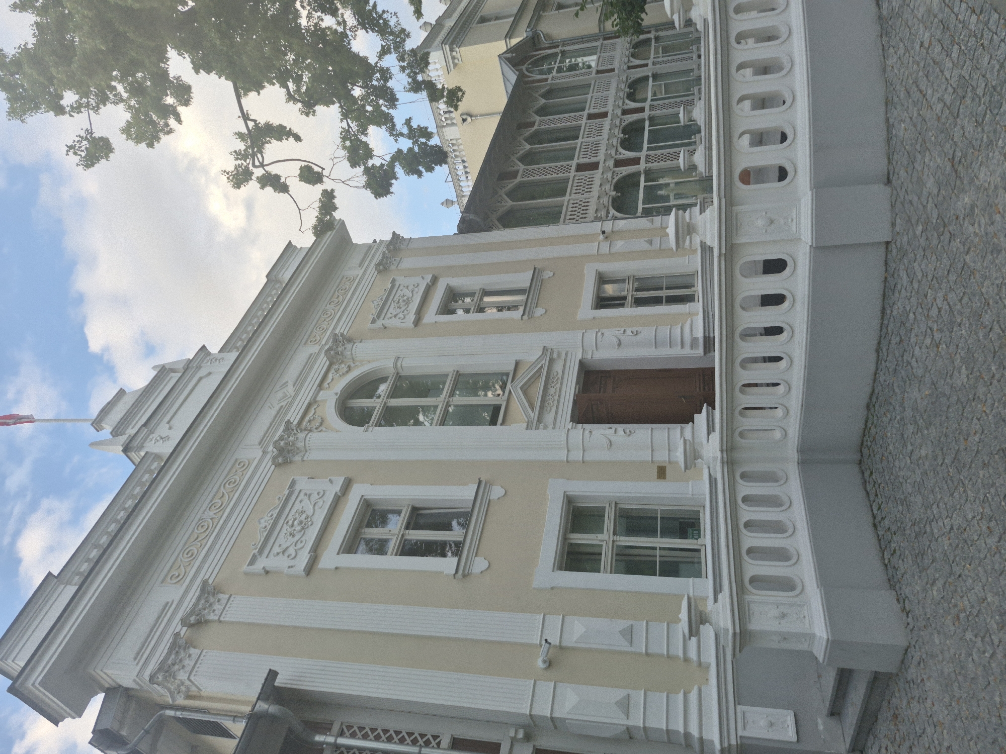 The image depicts the exterior of a pale yellow building with white ornate trim, situated in Gdańsk, Poland. The building has a classical architectural style with columns, decorative moldings, and arched windows. A white balustrade with oval openings runs along the front of the property, with cobblestone paving in front of it. To the right of the main building, another building with a similar color and style is visible, featuring a long, enclosed veranda with decorative white latticework. A tree with green leaves partially obscures the top right portion of the image. The sky above is a mix of blue and white clouds. The scene appears to be a street-level view of a historical building, possibly a museum, government office, or a grand residence.