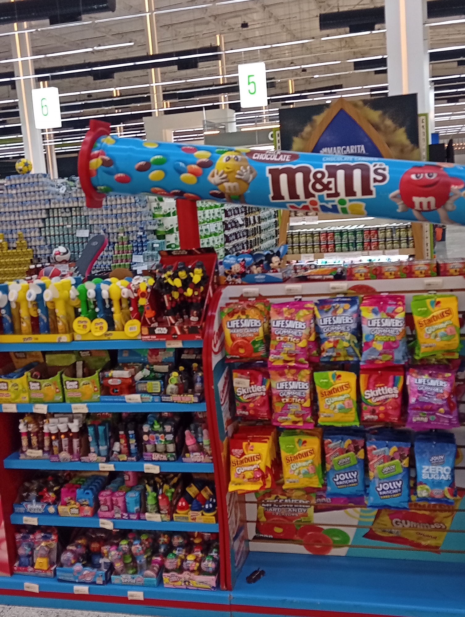 This image shows a candy display in a brightly lit store, possibly a supermarket, with shelving units filled with various candies and sweets. The top shelf features items like candy dispensers, while lower shelves showcase a variety of packaged candies like Life Savers, Skittles, Starburst, and Jolly Rancher gummies. A large M&M's Mini's promotional display sits above the shelves. The presence of aisle markers labeled "5" and "6" suggest a retail setting. The location is presumed to be Porlamar, Venezuela.