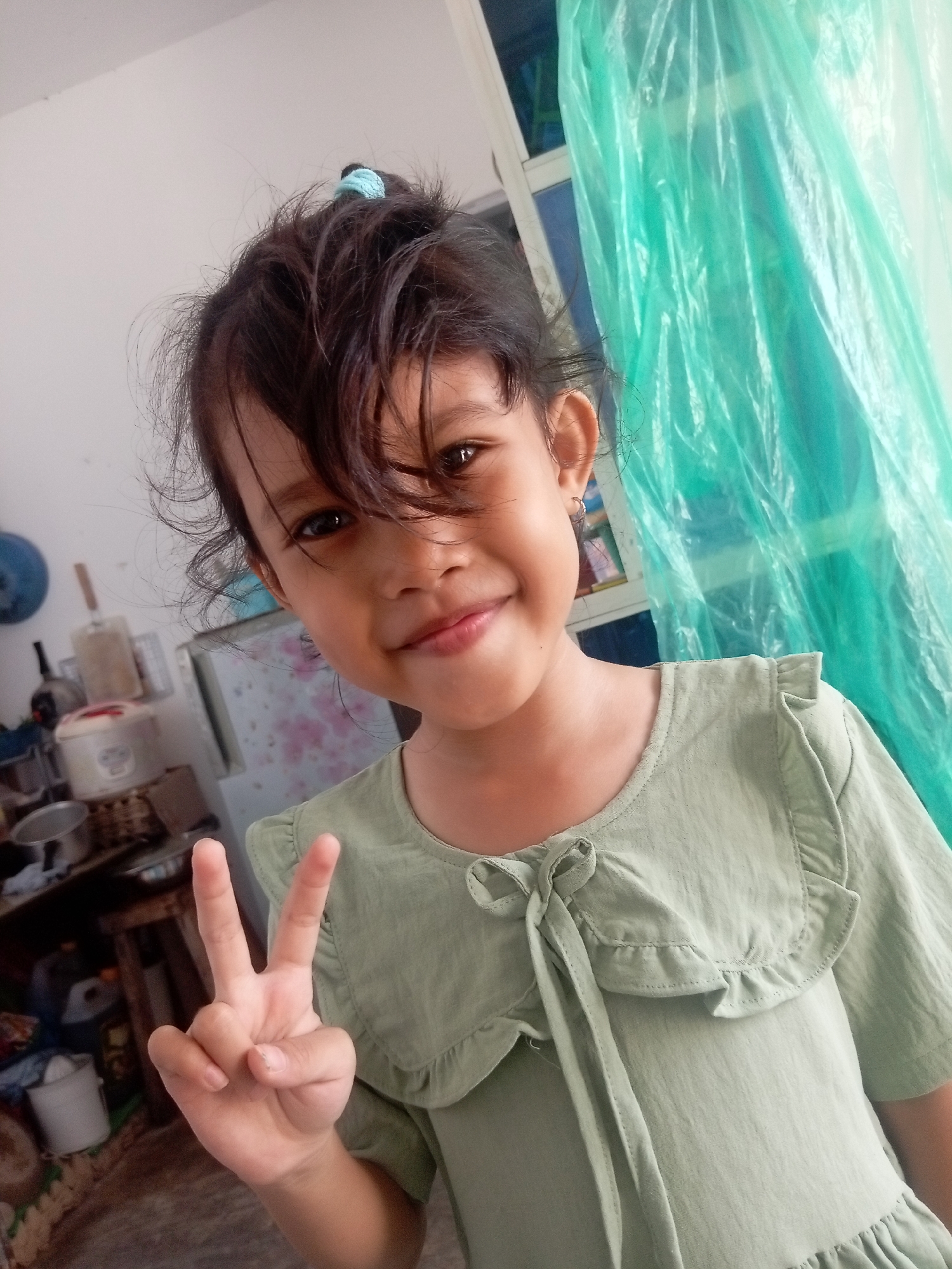 A young girl with dark hair tied in a messy bun with a blue scrunchie is smiling at the camera, making a peace sign with her left hand. She is wearing a light green dress with a ruffled collar and a bow detail. Her right hand is visible, resting on her body. The background is cluttered with household items, including a refrigerator, shelves, and various containers. A green plastic sheet is draped on the right side of the frame, partially obscuring a window. The lighting suggests an indoor setting, possibly a home in Batam, Indonesia, given the context.