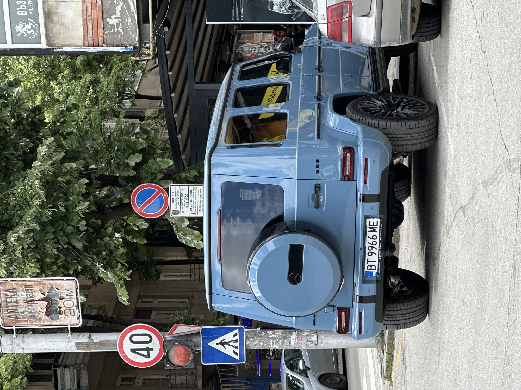 The image shows a light blue Mercedes-Benz G63 parked on a street in Veliko Tarnovo, Bulgaria. The car has a Bulgarian license plate "BT 9966 ME". Behind the car is a parked silver vehicle and a building with a black awning. In front of the car, there's a light pole with several signs: a "no parking" sign, a parking zone sign, a speed limit sign (40 km/h), a pedestrian crossing sign, and a faded sign indicating the distance to a pharmacy (20 meters). A traffic light shows a red light. Trees are visible in the background.