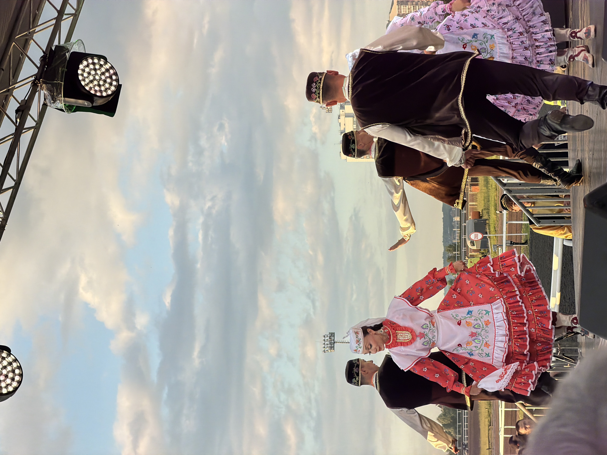 In the city of Unknown, Unknown, a group of performers in traditional attire are dancing on a raised stage. The foreground is dominated by the lower half of the performers, with their feet and legs visible. They are wearing dark boots, some with intricate designs. The performers are dressed in a variety of colorful, embroidered costumes. One female dancer in the center wears a red and white dress with elaborate floral embroidery and a white headpiece. To her left, a male dancer wears a dark vest over a white shirt and dark pants, along with a patterned cap. To the right, another male dancer is seen from the back, also in a dark vest and patterned cap, his movement suggesting he is dancing. Further to the right, a female dancer in a pink, patterned dress is partially visible. In the background, there are indistinct buildings and what appears to be a racetrack or stadium. The sky above is partly cloudy with a soft, natural light. Two stage lights are visible hanging from a metal structure above the stage.