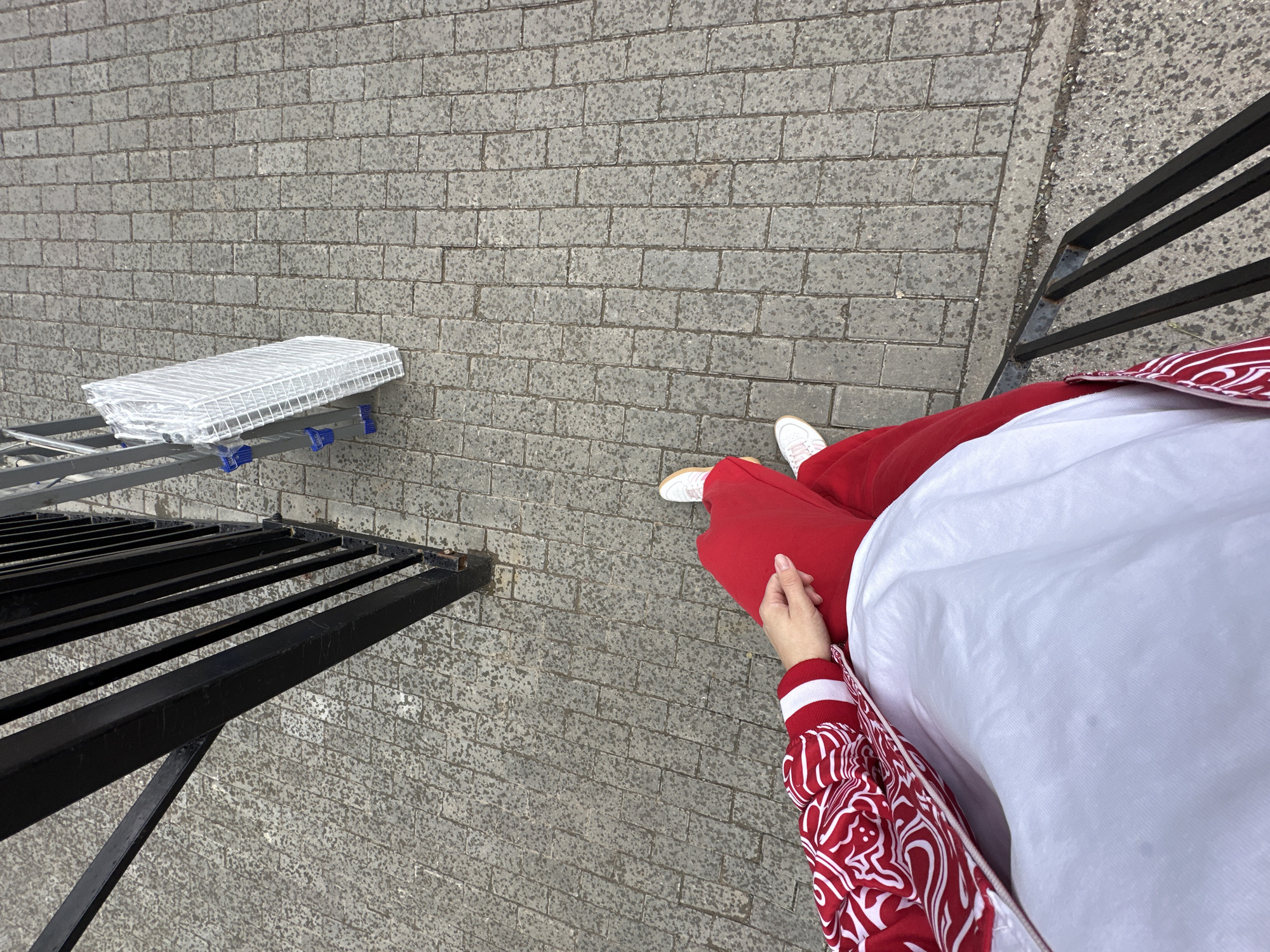 The image provides a top-down perspective of an outdoor paved area in Perm, Russia, featuring a person and various objects. The ground is composed of gray rectangular pavers or bricks, which have a mottled or speckled texture.
A person is visible on the right side of the frame, dressed in red pants, a white long-sleeved shirt, and a red and white patterned jacket featuring a dynamic, swirling design. White sneakers are visible on their feet, and one hand rests on their thigh.
On the left side of the image, a metallic structure, possibly a ladder or part of a display stand, supports several white wire grids that are individually wrapped in clear plastic. Further to the left and along the right edge, black metal railings or frames are partially visible, suggesting an architectural boundary or structure.
The scene depicts a static moment, with the person standing. The lighting suggests it is daytime. No specific emotions are discernible, and no text is visible within the image.