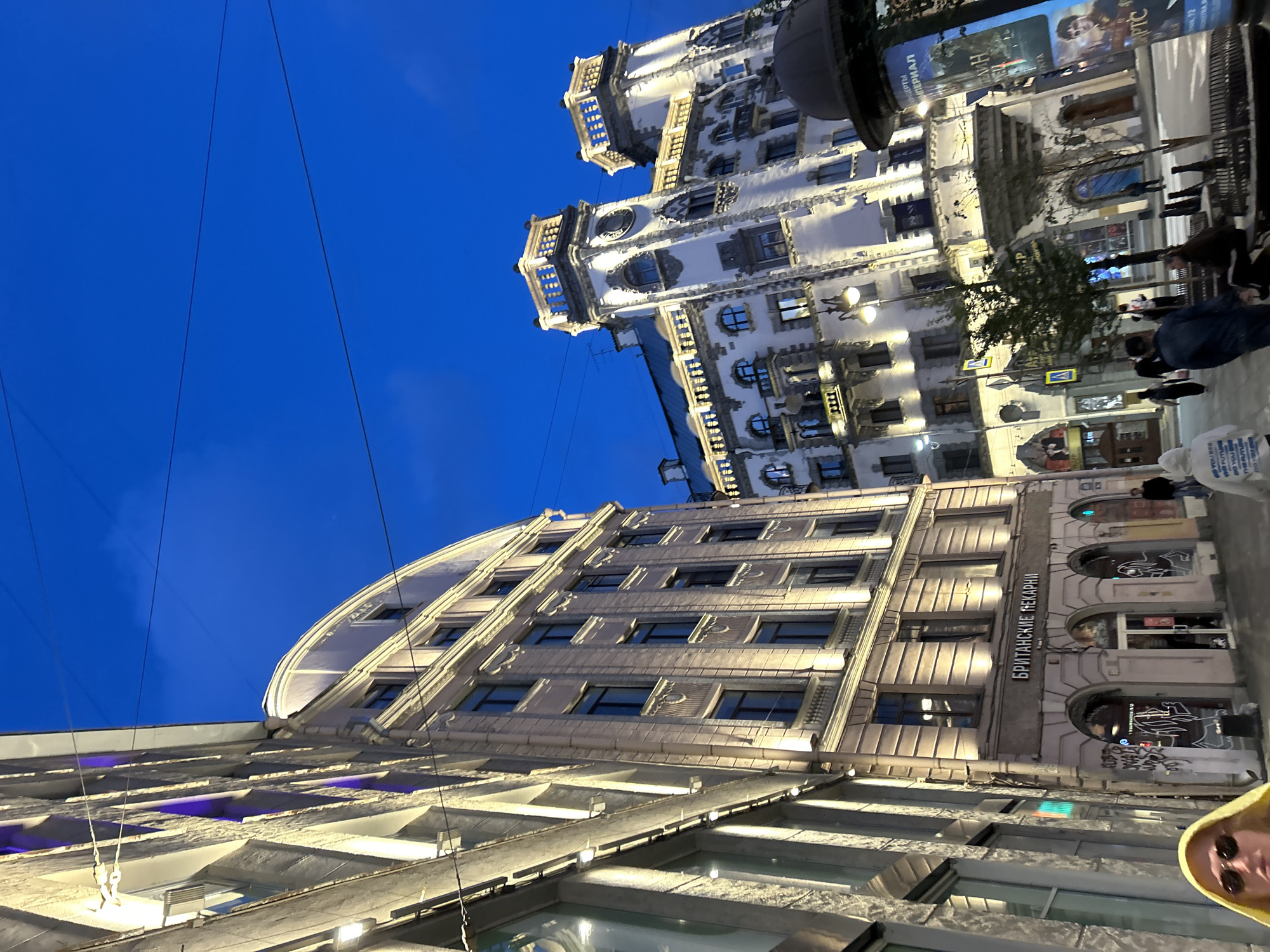 The media file captures an upward-angle view of several buildings at dusk. The sky is a deep, clear blue, indicating it is likely evening. The buildings are illuminated, with lights visible in many windows, suggesting the activity within.
In the foreground, a modern building with large windows and exterior lighting is visible. The building is multi-story, with a ribbed or textured facade. Some sections of this building are lit with purple light. Further back, a more ornate building with a white facade, intricate architectural details, balconies, and arched windows is prominent. This building appears to be older and is also well-lit, highlighting its decorative features. The perspective shows these buildings on a street, as indicated by the presence of people walking, streetlights, and what appears to be a lamppost.
One notable detail is the text "БРИТАНСКИЕ ПЕКАРНИ" visible on the facade of one of the buildings, which translates to "British Bakeries" in Russian. This suggests a potential location in a Russian-speaking country.
The scene conveys a sense of urban life at the end of the day, with the buildings standing out against the twilight sky. The lighting creates a warm and inviting atmosphere. A person wearing sunglasses and a yellow hoodie is partially visible in the bottom right corner, looking towards the camera, adding a human element to the urban landscape. The overall impression is one of architectural beauty and evening activity in a city.