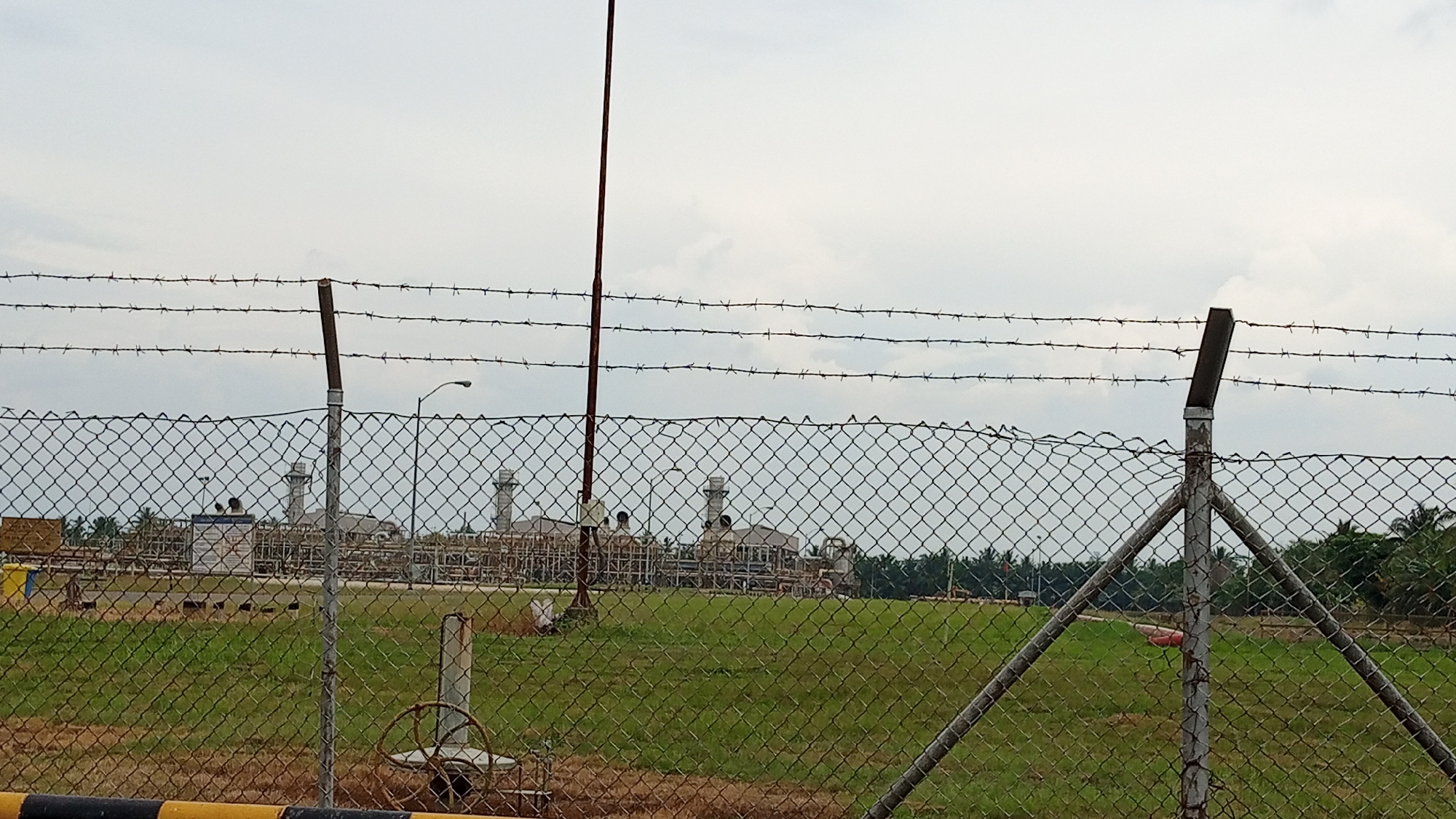 The image depicts an industrial facility, likely a power plant, viewed through a chain-link fence topped with barbed wire. In the foreground, the fence is prominent, with a metal post and diagonal supports visible. Beyond the fence, a grassy field stretches towards the industrial complex. The complex itself features several large structures, including tall smokestacks or chimneys, and numerous pipes and smaller buildings. A lamppost is also visible within the facility grounds. The sky is overcast with clouds. The overall scene suggests a secure, operational industrial area, possibly in Glokaron, Indonesia, given the context. There are no visible people in the image.