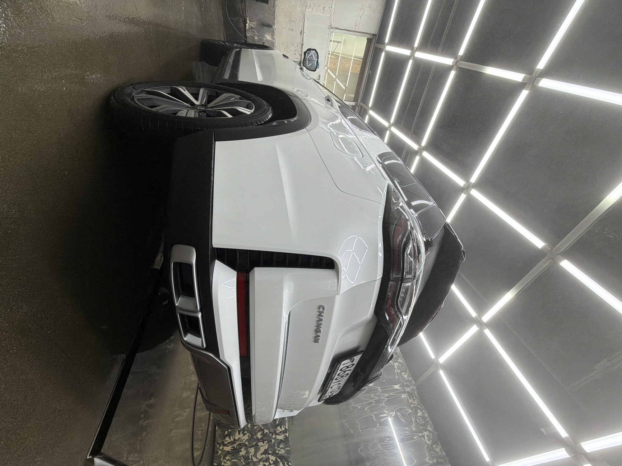 A white Changan car is parked inside a detailing or car wash studio. The vehicle is oriented with its rear end facing the viewer, showcasing the taillights, bumper, and license plate. The word "CHANGAN" is clearly visible on the lower portion of the car's rear. The environment features a bright, evenly lit space with rows of linear lights creating a grid pattern on the ceiling and walls. The floor appears to be wet. The license plate, "T868TC124", suggests the car is registered in the Krasnoyarsk region of Russia.