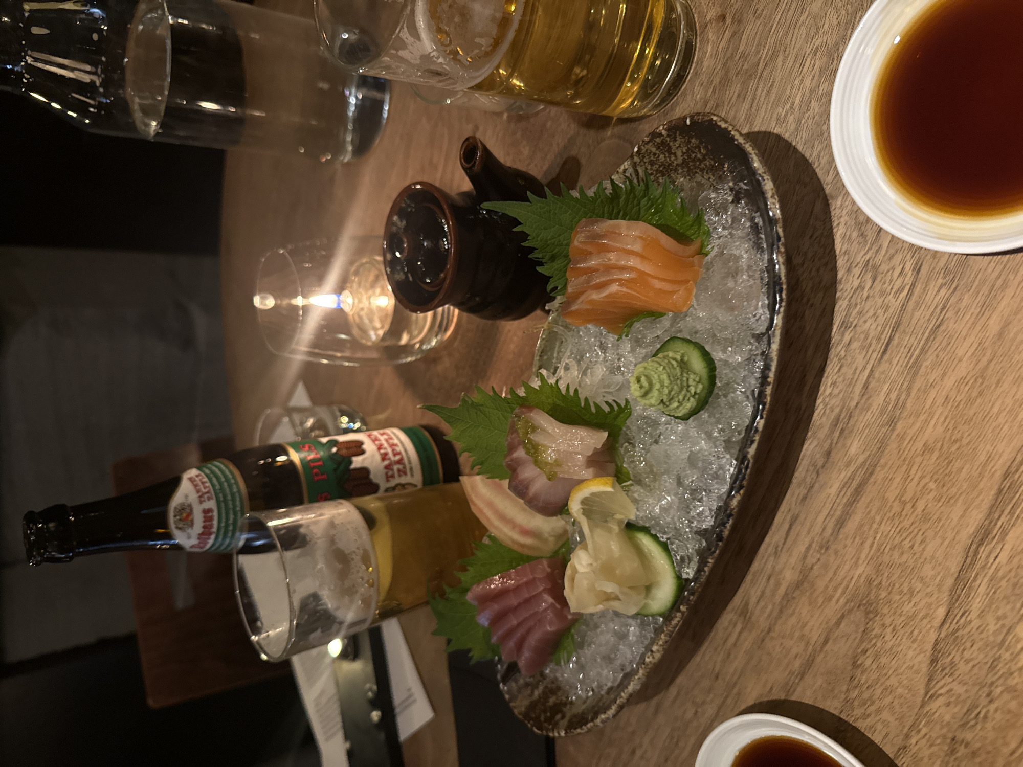 The image displays a dining scene, likely in a restaurant or bar in London, presented from an overhead perspective. The setting suggests an evening ambiance, illuminated by a flickering candle in a clear glass holder.
The primary subject is an oval-shaped ceramic platter of sashimi resting on a bed of crushed ice. The platter features several varieties of fish, including slices of salmon, tuna, and white fish, artfully arranged with garnishes such as fresh green shiso leaves, thinly sliced cucumber, a wedge of lemon, pickled ginger, and a dollop of green wasabi paste.
Accompanying the meal are drinks and condiments. A bottle of beer, clearly labeled "Rothaus TANNENZÄPFLE Pils," stands upright, beside which a glass filled with golden beer is visible. Another glass containing beer and an empty glass are also present. A small, dark ceramic pot with a spout, potentially for pouring soy sauce or sake, sits near the sashimi. A small white bowl holds a pool of dark soy sauce. The entire arrangement is set on a warm-toned wooden table. No people are visible in the photograph.