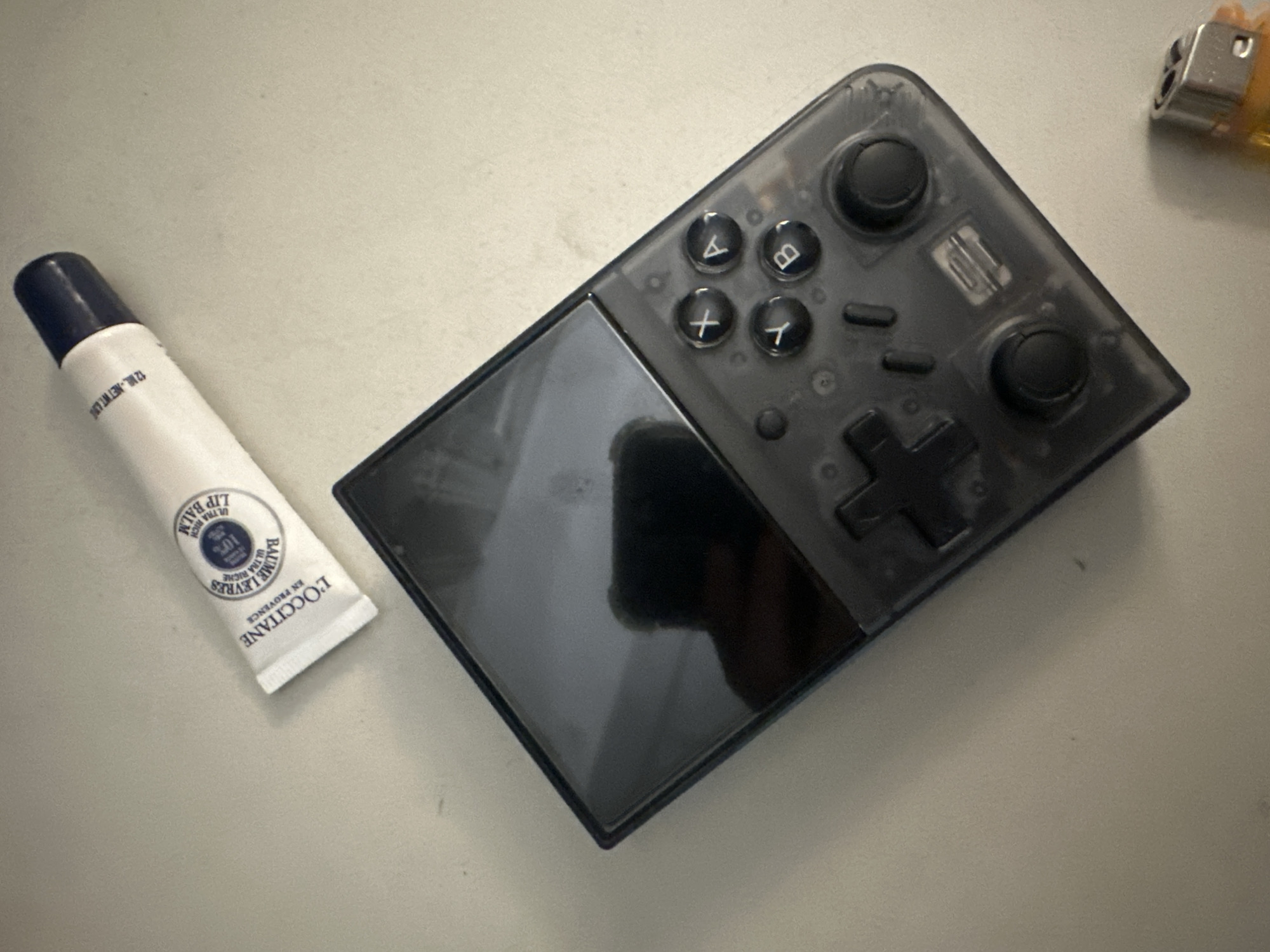 An overhead shot displays three personal items resting on a light grey, textured surface, likely a table or desk, in Buenos Aires, Argentina.
To the left, a white tube of lip balm with a dark blue screw cap is visible. The text on the tube identifies it as "L'OCCITANE EN PROVENCE BAUME LEVRES ULTRA RICHE" and "LIP BALM ULTRA RICH," indicating a "10%" enrichment. The volume is specified as "12 ML. NET WT. L".
Dominating the right side of the frame is a compact handheld gaming console with a translucent dark grey casing, revealing some of its internal components. The control layout includes four prominent face buttons marked 'A', 'B', 'X', and 'Y', a black D-pad, and two black analog sticks. Several smaller function buttons are also present. The console's screen is dark, reflecting the surrounding environment.
In the upper right corner, partially visible, is the metallic top and a sliver of the yellow body of a lighter. The image is a static composition of these objects; no specific activity is taking place.