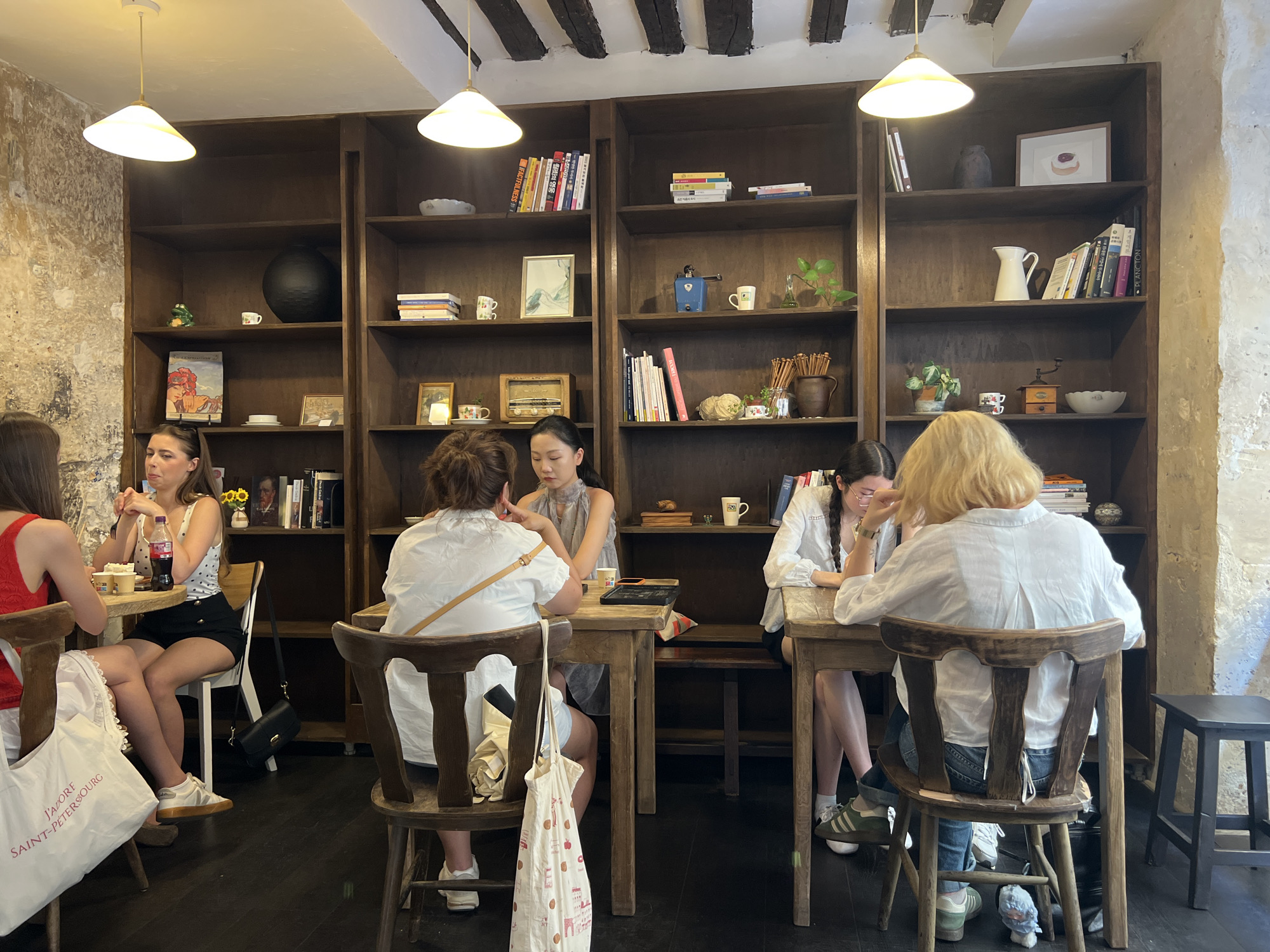 This image depicts the interior of a cafe in Paris, France, likely an establishment designed with vintage or rustic decor. The scene is illuminated by warm light from pendant lamps hanging from a beamed ceiling.
In the foreground, four women are seated at tables. On the left, a woman in a red top and white skirt is visible from the chest down, and a large white tote bag with French text, "J'ADORE SAINT-PETERSBOURG," is placed on her chair. Next to her, a woman wearing a polka dot top and black shorts is seated, holding a fork and looking at her food. A bottle of Coca-Cola and a cup are on the table.
In the center, a woman in a grey dress is seated, her back to the camera, facing a table with a black tray. To her right, another woman in a white top with braided hair is seated at a table, looking down.
To the right, the back of a woman with blonde hair wearing a white shirt and blue jeans is visible. She is seated on a wooden chair facing a wooden table. Her legs are crossed, and she is wearing green and white sneakers.
The background features tall, dark wooden bookshelves filled with books, decorative items, and some ceramic pieces. Notable items on the shelves include a large dark vase, small ceramic figurines, a vintage radio, and various books with colorful spines. The walls appear to be textured, possibly stone or stucco, with some visible wear and tear. The flooring is dark wood. The overall atmosphere suggests a relaxed, indoor setting within a cafe.