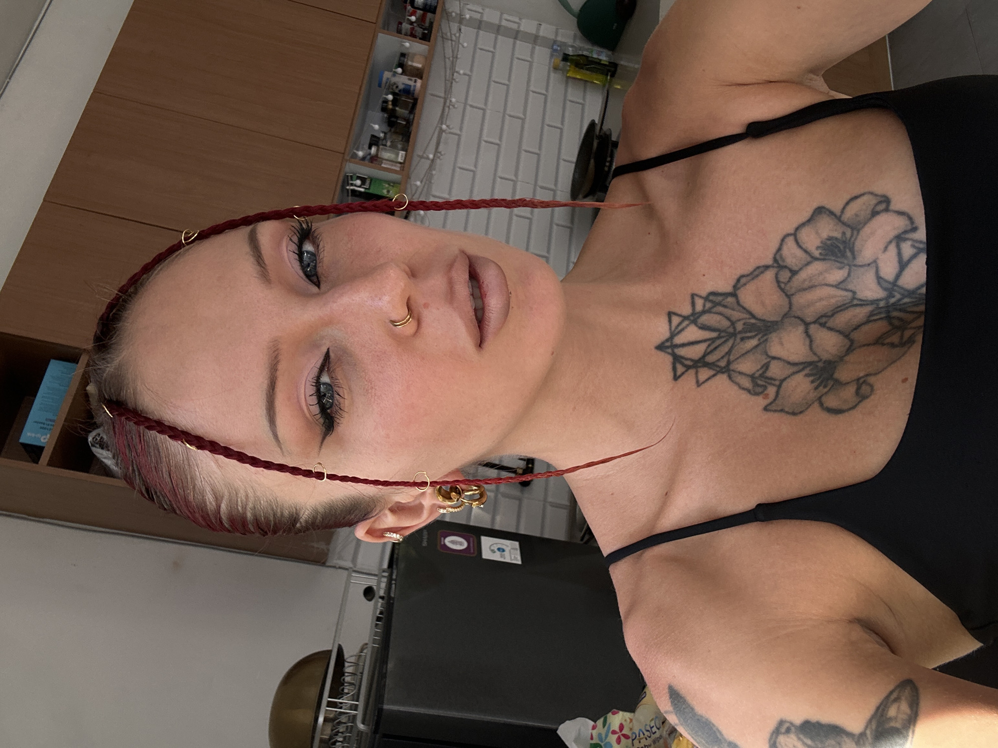 A woman with red braided hair and various piercings poses for a selfie in a kitchen. The woman has makeup on, including eyeliner and lipstick. She is wearing a black tank top. A tattoo of flowers and geometric shapes is visible on her chest. In the background, kitchen cabinets and appliances are visible. The scene appears to be indoors, likely in a residential kitchen.