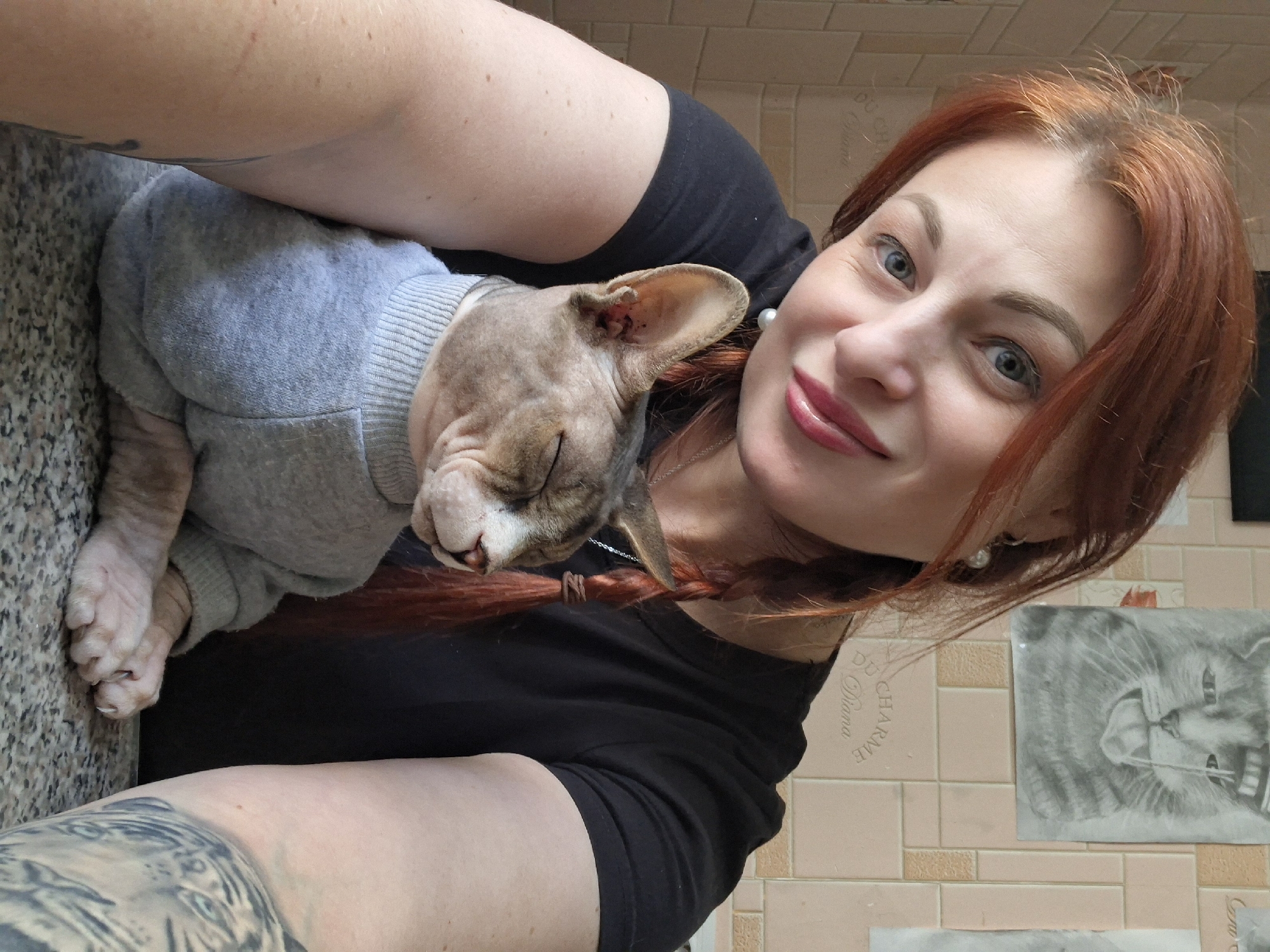 In an indoor setting, a woman with reddish-brown hair styled in braids and bright blue eyes smiles at the camera. She is wearing a black t-shirt and a pearl earring. She is holding a hairless cat, which is wearing a light grey sweater. The cat's eyes are closed, and it appears relaxed. Behind the woman, a tiled wall is visible, with a drawing of a cat in the upper portion. The scene suggests a casual, personal moment captured at home.