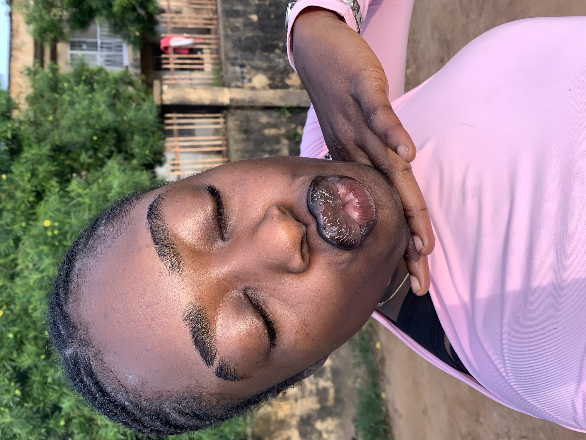 A close-up, angled shot shows a young Black woman with her eyes closed and her lips puckered, wearing a pink long-sleeved shirt and black undergarment. Her hand is resting on her chin. The background includes lush green trees, a fence, a building, and a dirt path. The setting appears to be outdoors in a residential area, likely in Rafin Par, Nigeria, given the architectural style and vegetation common to the region.
