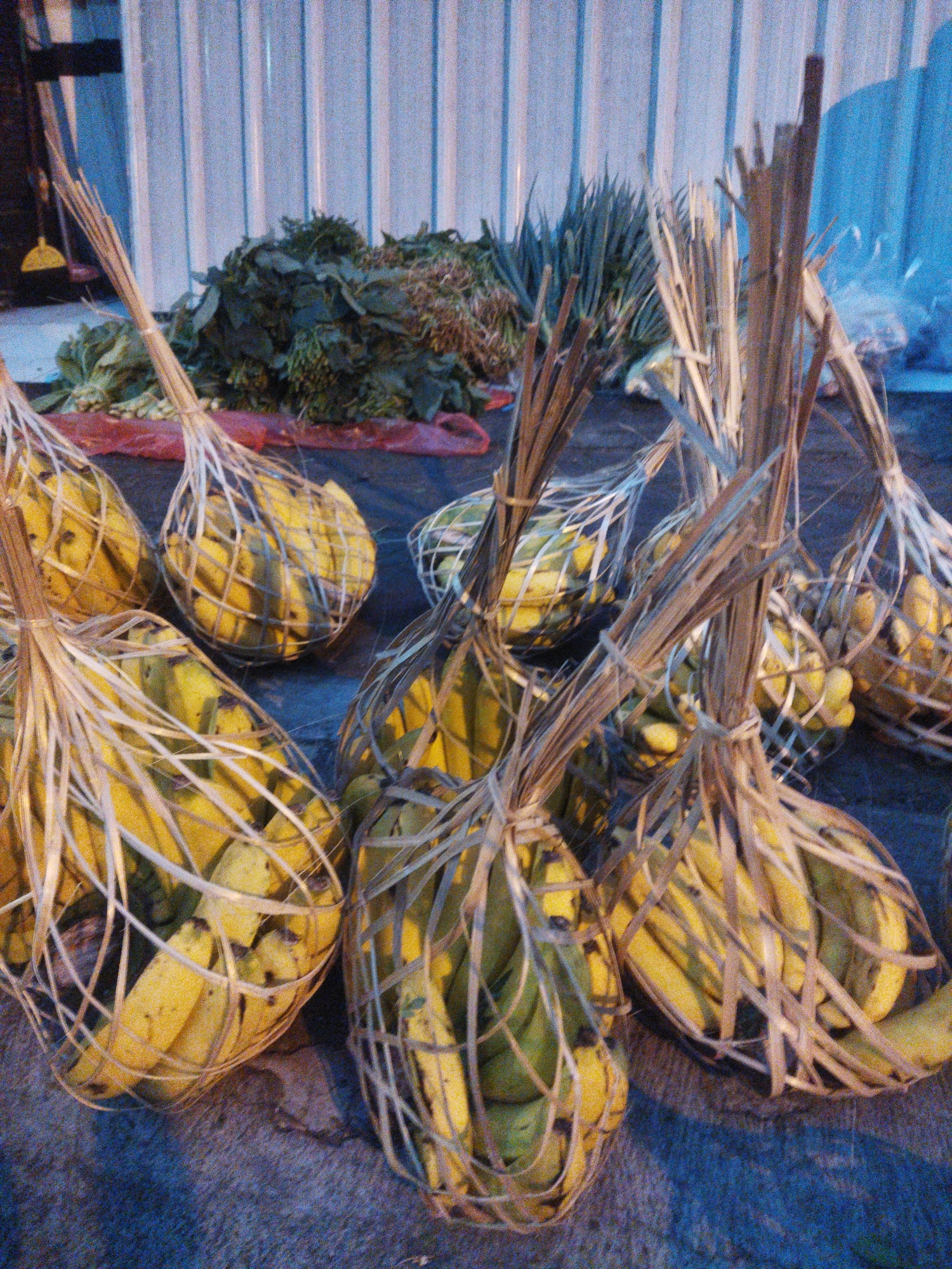This is a still photograph of a collection of bunches of bananas, each wrapped in a woven straw basket, arranged on a dark, textured surface. The bananas are predominantly yellow, with some green hues visible. Behind the bananas, a pile of leafy green vegetables is present, and further back, a corrugated metal wall or fence is visible. The lighting suggests it may be dusk or early morning. The scene is indicative of a market stall or a vendor preparing produce for sale. The presence of bananas bundled in straw baskets is a common sight in Indonesian markets, and the leafy greens suggest a connection to local agriculture.