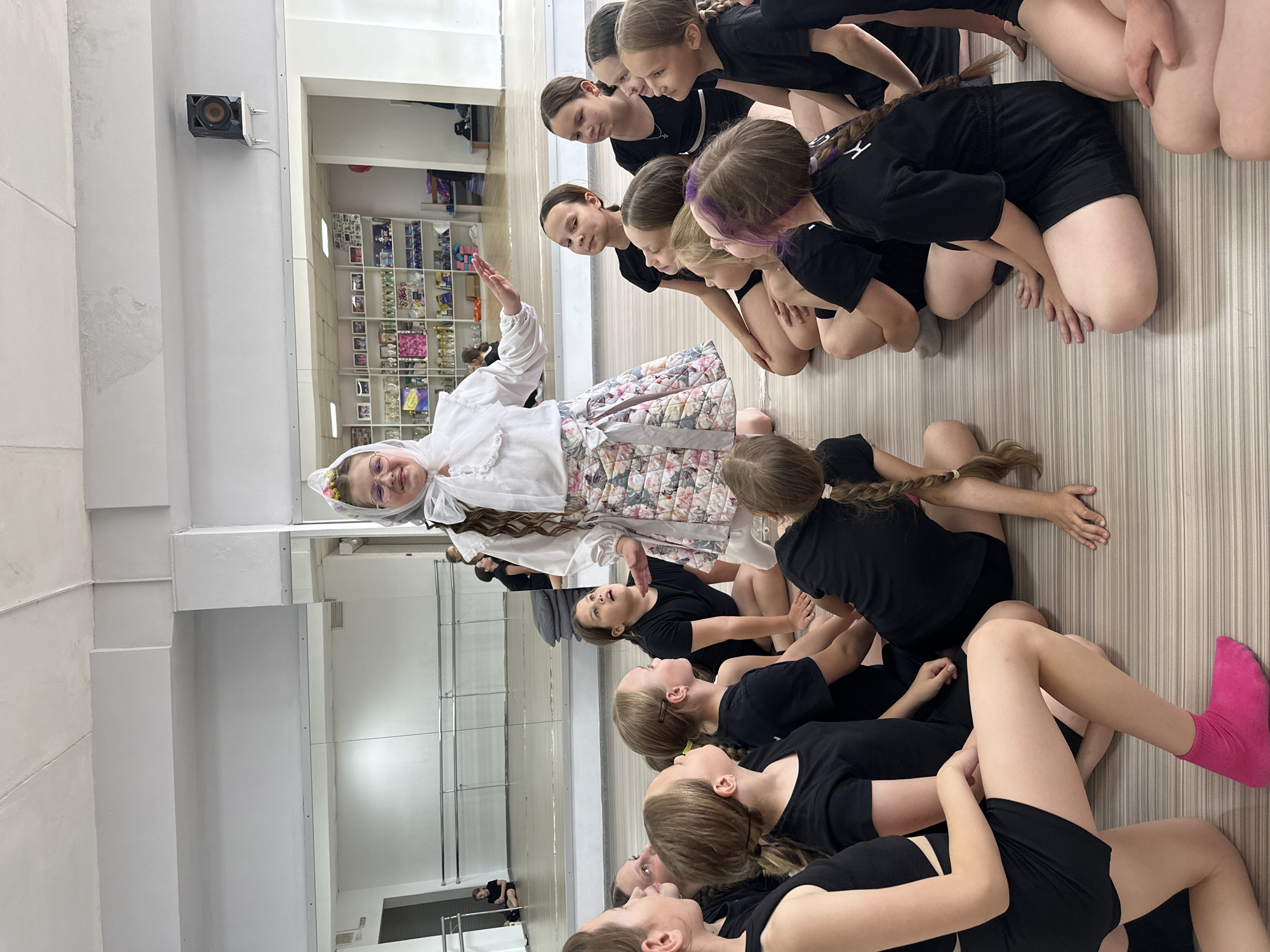 A group of young girls are gathered in a dance studio. Some of the girls are seated on the floor facing a woman in traditional Russian clothing. The woman is standing and gesturing with one hand. The girls are wearing black dance attire, and the woman is wearing a white blouse, a floral skirt, and a white headscarf. The dance studio has light-colored walls and a striped floor. The walls are decorated with mirrors and shelves. A small speaker is attached to the wall. The scene appears to be a dance class or rehearsal, possibly related to Russian folk dance. The location in the city of Votkinsk, Russia, is not verifiable from the given image.
