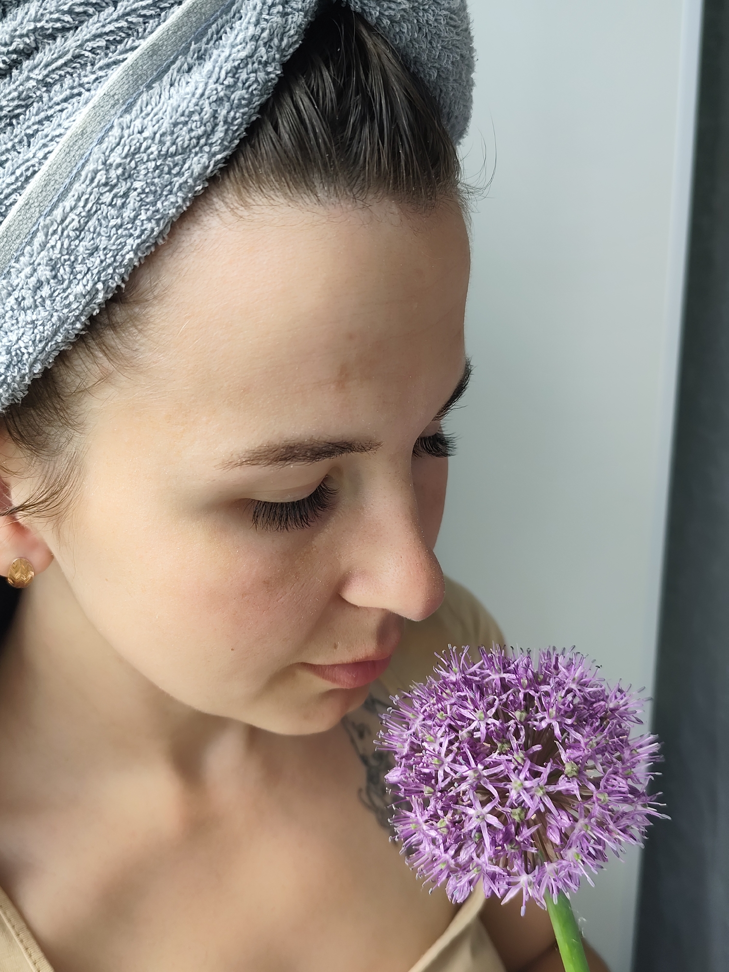 A woman with a gray towel wrapped around her head is holding a purple allium flower near her face, looking down at it. She has dark hair, light skin, and noticeable eyelash extensions. She is wearing a light-colored top and a gold stud earring is visible. A small tattoo is visible on her upper chest/shoulder area. The background is a white wall with a dark gray curtain to the right. There is no indication of the location being in the city of Фоки, Russia.
