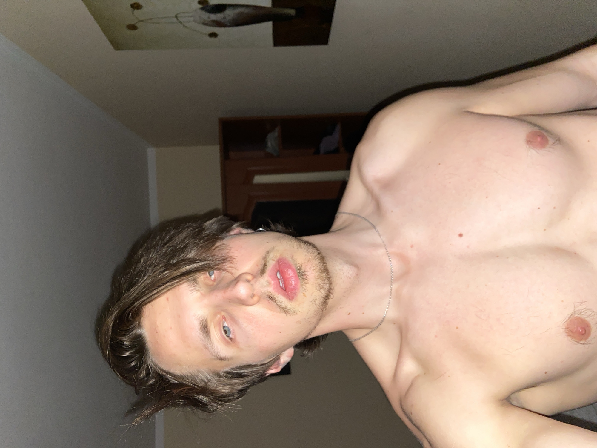 A shirtless man with light skin, brown hair, and blue eyes is lying down. He is wearing a silver chain around his neck. The background shows a wall, a piece of artwork hanging on the wall, and a dark wood dresser. The location, Šušanj, Montenegro, cannot be confirmed from the image.