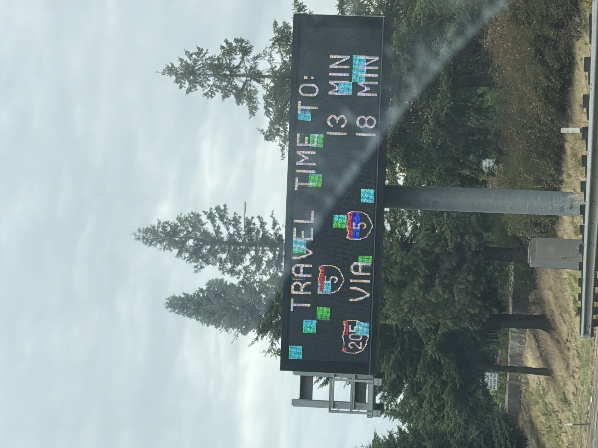 The media file is a vertical, slightly tilted, outdoor photograph of a digital road sign on a cloudy day. The sign displays travel information. On the left side, it reads "TRAVEL TIME TO:" in large, colorful, pixelated letters. To the right of this, it shows a travel time of "13 MIN" and "18 MIN", with the number "5" displayed prominently, indicating a route. Below this, the sign shows "205 VIA 5", suggesting a specific route or highway. There is also some fragmented text at the bottom left that is difficult to read completely. The background consists of dense green trees and a cloudy, overcast sky. The sign itself is mounted on a metal pole structure. In the distance, there are more trees and a hint of dry, brown grass on the side of the road. A small white sign is visible among the trees in the background, but the text is too small to read. This appears to be a highway scene in Beaverton, United States of America. The overall atmosphere is muted due to the overcast weather.
