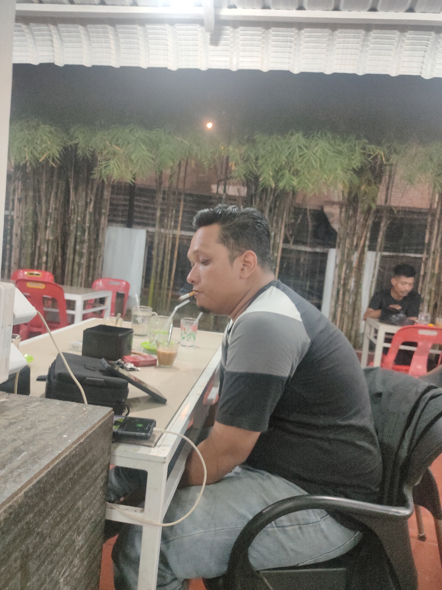A man wearing a gray and black shirt, and light blue jeans sits at a white table in an outdoor setting. The man is smoking a cigarette. He is seated in a black chair. On the table are a black bag, a phone, glassware, and other items. Behind him, bamboo plants are visible, and another man is seated at a table in the background. The location is likely in Lhokseumawe, Indonesia. The scene appears to be a casual outdoor café or restaurant setting.