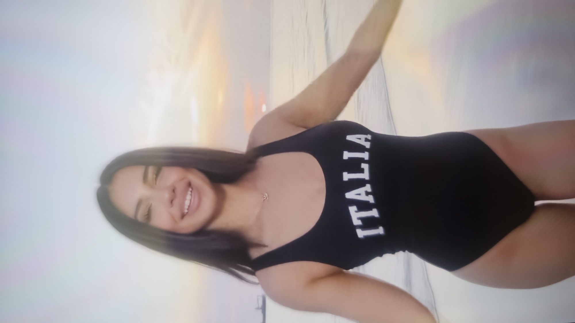A young adult female is positioned on a sandy beach at sunset. She has long, dark, straight hair, parted centrally, and a broad smile revealing her teeth. She is wearing a black one-piece swimsuit with the word "ITALIA" printed in white block letters across the chest. A delicate gold-toned necklace with a small pendant is visible around her neck. Her arms are slightly extended outwards from her body. The background features a sunset sky displaying a gradient of warm colors, transitioning from orange, pink, and yellow at the horizon to lighter blue and purple higher up. The sun is partially visible on the horizon, casting a warm glow. Calm water with gentle waves is present, breaking onto the sandy shoreline. In the distant left, two dark, indistinct silhouettes, consistent with ships or structures, are visible on the water's horizon.