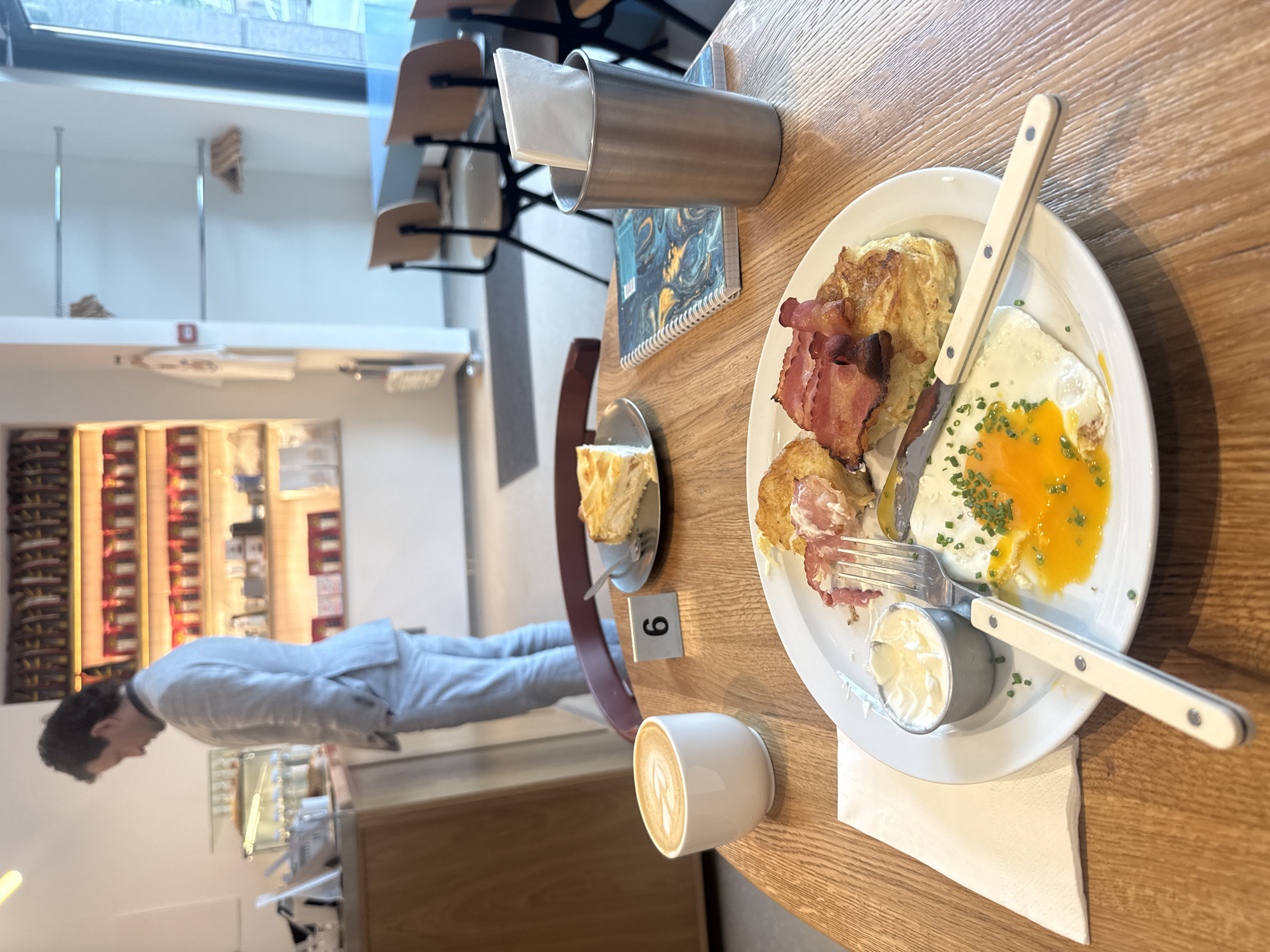 An indoor scene shows a table in a cafe with a breakfast meal and a cup of coffee. The table is made of light-colored wood. On the table, a white plate holds a fried egg with a runny yolk, sprinkled with chives, two strips of bacon, and a piece of pastry. A small metal ramekin of sauce sits next to the egg. A white napkin is folded to the left of the plate, and a fork and knife with cream-colored handles are placed on the plate. A white ceramic cup with latte art sits to the left of the plate. A small metal container holding napkins is to the right of the plate, behind a spiral-bound notebook with a blue and gold marbled cover. A table number "6" is visible on a black stand behind the coffee cup. In the background, a person in a light gray suit is standing with their hands in their pockets, facing away from the camera and towards a counter. Behind the person, shelves are stocked with numerous small red and gold packaged items. In the far background, there are tables and chairs.