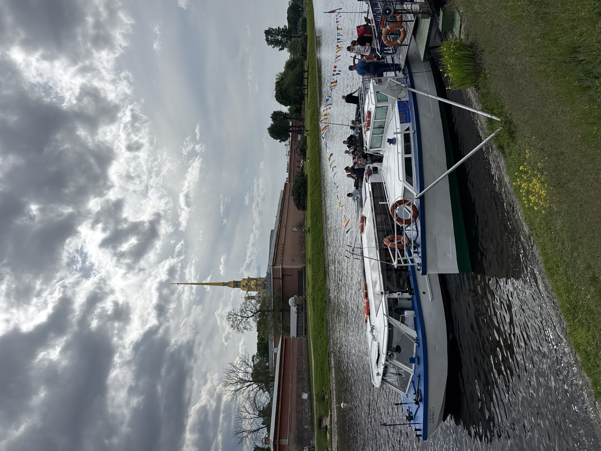 This photograph depicts a scenic river or canal view in Saint Petersburg, Russia, showcasing two tour boats in the foreground and the historic Peter and Paul Fortress and Cathedral in the background.
In the right foreground, two white boats, likely passenger vessels for river excursions, are positioned on the water. Several people, appearing to be tourists, are visible on their decks, either standing or seated, observing the surroundings. The boats feature blue and green accents on their hulls, and orange life preservers are mounted on their railings. Yellow flowers dot the grassy bank that borders the water on the right.
Across the waterway, the distinctive red-brick walls of the Peter and Paul Fortress are prominently displayed, stretching along the opposite bank. Beyond the fortress walls, the magnificent golden spire of the Peter and Paul Cathedral rises high into the sky, topped with a cross. Trees with varying degrees of foliage are visible within and around the fortress grounds. Decorative strings of international maritime signal flags are strung along the riverbank on the fortress side, adding a festive element to the scene.
The weather appears overcast with a dynamic sky featuring bright, textured clouds interspersed with darker grey areas, suggesting a cloudy but bright daytime. The overall mood is calm, indicative of a leisurely boat tour.
Visible text on the hull of the lower boat reads: "C3 15-41 KOPBET-1".