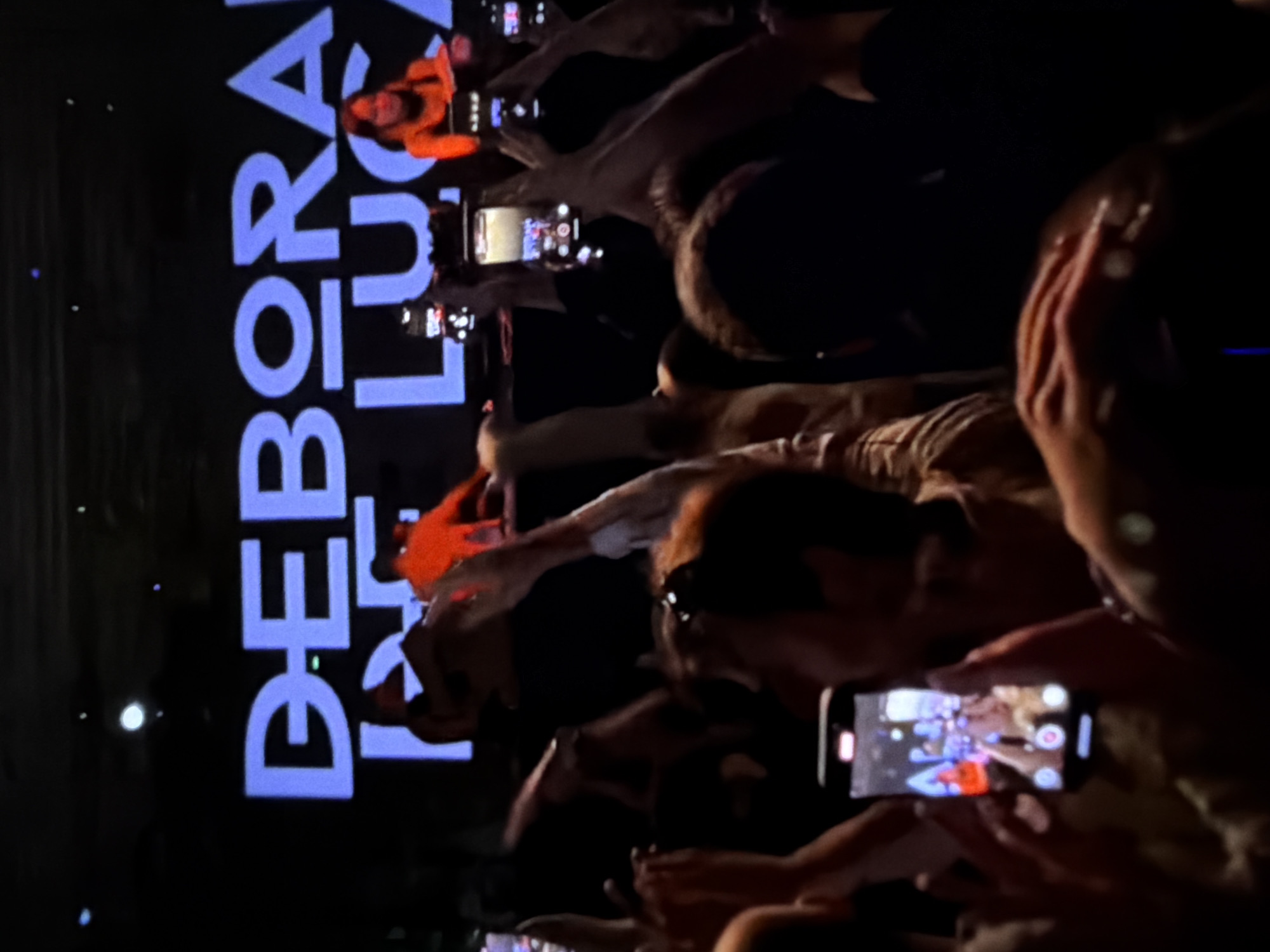 Here is a description of the image:
The image captures a concert scene, likely indoors given the darkness. The words "DEBORA LUC..." are prominently displayed in vertical text, possibly on a screen or backdrop. A crowd of people is visible, many holding up their phones to record or take pictures of the stage. Several phones are distinctly visible, showing live video feeds. The lighting is focused on the stage, with an orange glow illuminating some figures. The environment seems energetic and crowded, typical of a live music event.
