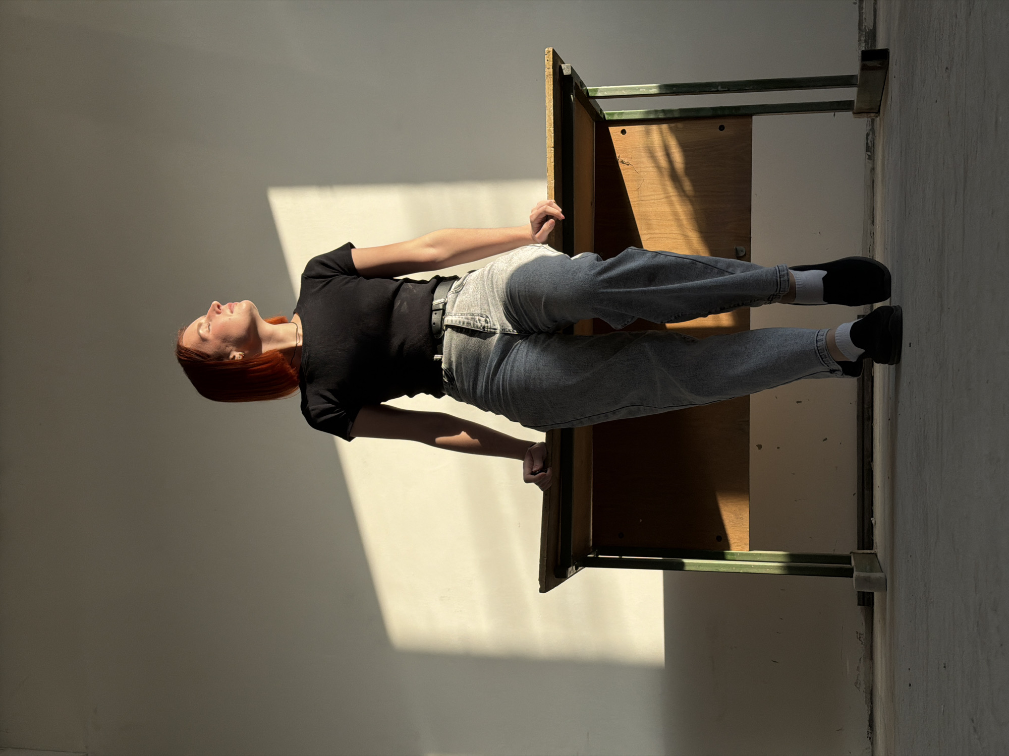 A young adult female with short, red hair, dressed in a black ribbed t-shirt, light grey denim jeans, a black belt, white ankle socks, and black slip-on shoes, is seated atop a worn wooden desk with a dark green metal frame. Her hands rest on the desk's surface on either side of her. Her head is tilted slightly back, and her face, with closed or squinting eyes, is turned towards a bright patch of sunlight illuminating the plain, light-colored wall behind her. The desk's feet are propped up on small concrete blocks. The environment is an indoor room with bare, light-colored walls and a concrete floor. Strong, directional sunlight casts sharp shadows across the wall and floor from an unseen source to the left. The specific location within the city of Chaikovsky, Russia, is not discernible from the visual information provided.