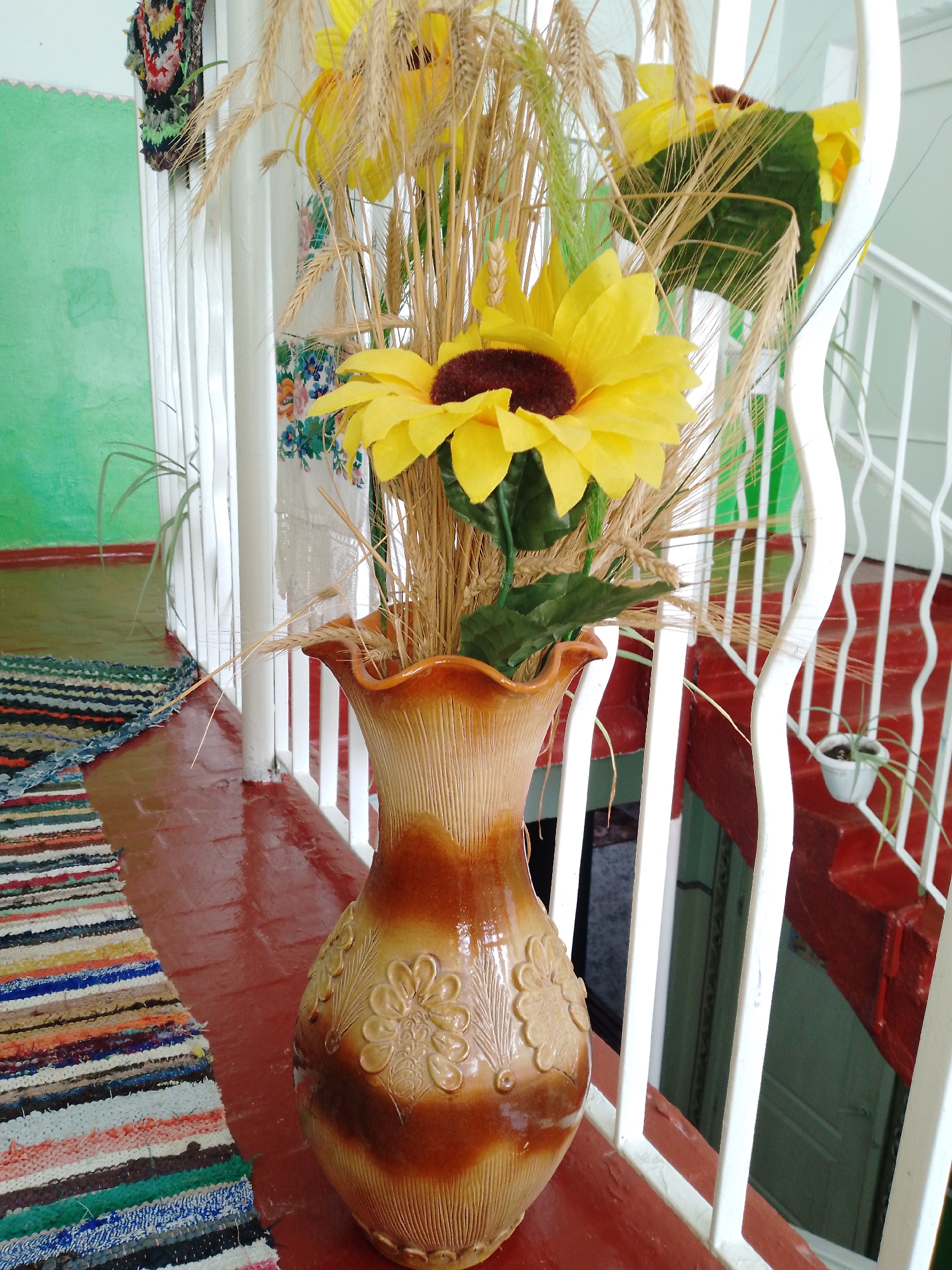 An indoor scene in Ustyvytsia, Ukraine, centers on a decorative ceramic vase holding a bouquet of artificial sunflowers and dried wheat stalks. The vase, positioned on a reddish-brown ledge, is two-toned with a glazed tan top and a darker brown lower section featuring embossed floral patterns. The sunflowers have bright yellow petals and dark brown centers, complemented by green artificial leaves. The dried wheat stalks stand tall, intermingled with the sunflowers, giving the arrangement an autumnal or harvest feel.
To the left of the vase, a colorful, striped rag rug with shades of blue, red, green, brown, and white lies on the reddish-brown floor. The floor extends towards a light green painted wall in the background, where a small green plant with long, thin leaves is visible against the wall. In the upper left corner, a piece of vibrant, multicolored woven textile art hangs on the green wall.
A white wooden or metal banister/railing runs vertically, separating the main floor level from a lower area. Through the railing, glimpses of a white fabric hanging, possibly embroidered with colorful floral patterns, can be seen behind the vase. To the right of the vase and railing, a set of red-painted stairs descends. A small white potted plant with green foliage rests on one of the red steps. The overall lighting suggests it is daytime. No people or text are visible in the image. The elements like the sunflowers, wheat, and traditional-looking rug evoke a sense of Ukrainian folk decor and a humble, lived-in domestic setting.