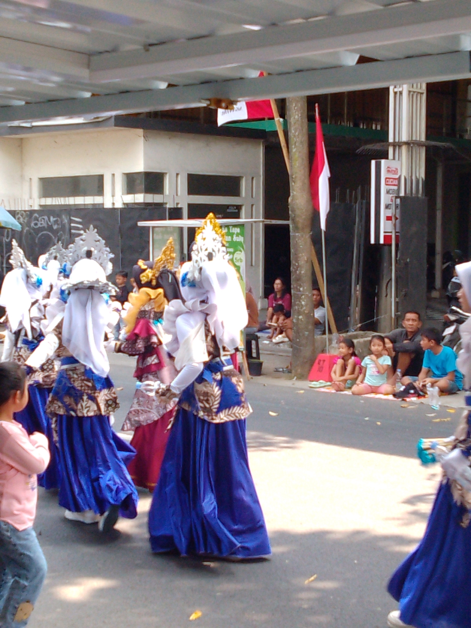 In the city of Trenggalek, Indonesia, a cultural parade is underway. Several performers, adorned in elaborate blue and white costumes with intricate gold and silver headdresses, are walking down a paved street. They are likely participating in a local festival or celebration. Spectators, including children and adults, are seated on the side of the road, observing the procession. Some are sitting on the pavement, while others are on raised steps. The environment suggests an outdoor event, with buildings and trees visible in the background. The mood appears festive and engaging, with the colorful costumes and the presence of an audience.