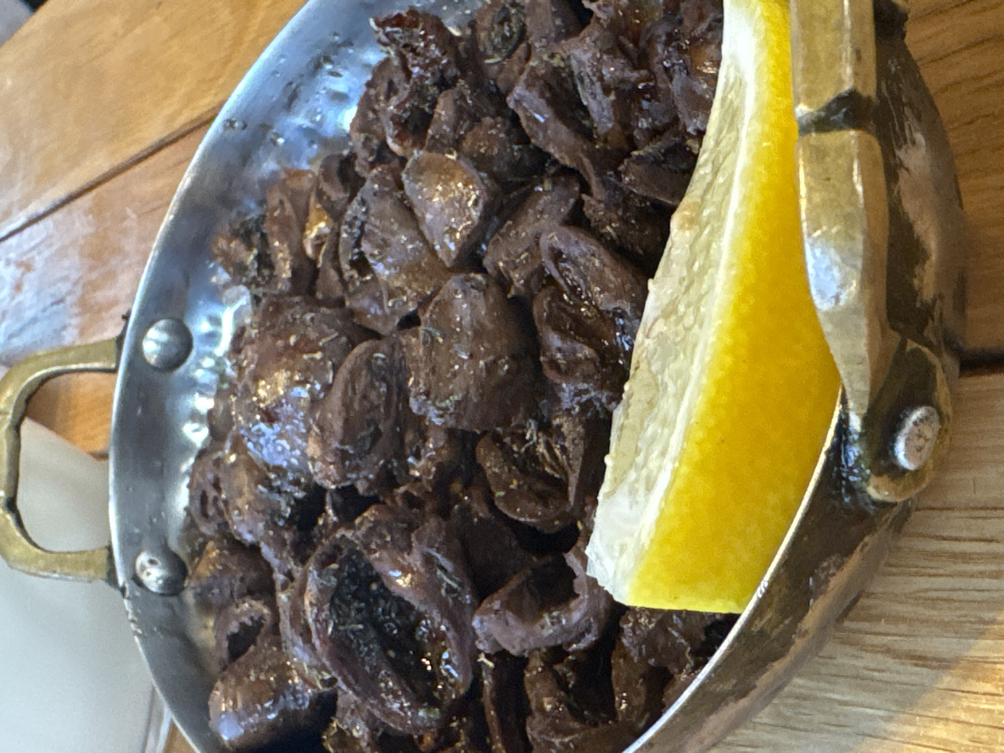 A close-up shot shows a metallic bowl filled with dark, stewed food, possibly olives, seasoned with herbs. A wedge of bright yellow lemon rests on the edge of the bowl, in the foreground. The bowl is set on a wooden surface. The image is taken from a slightly elevated angle. The background is blurred, with hints of white and warm tones. The scene appears to be from a restaurant in Primorsko, Bulgaria, suggesting a typical meal presentation.