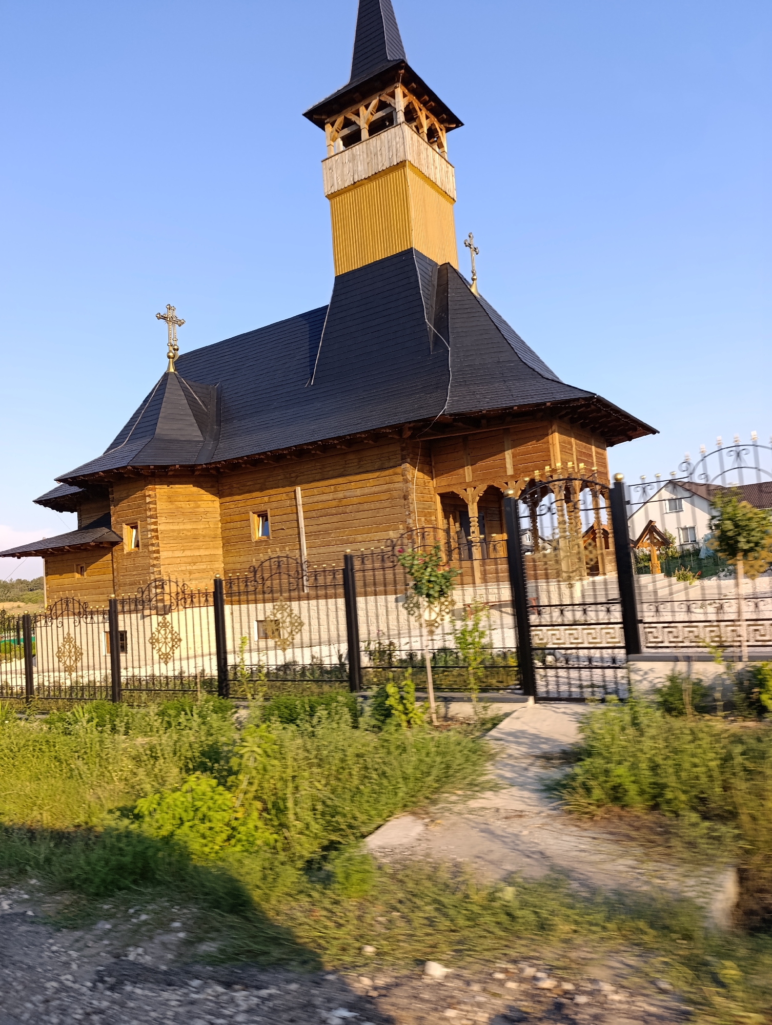 An outdoor, medium-shot, eye-level, vertical, full color photograph shows a wooden Orthodox church with a tall, slender, golden bell tower topped with a black spire. The church's roof is black and multi-tiered. The building is constructed of light-colored wooden planks. A golden cross sits atop the bell tower and another on the main roof. A black metal fence with decorative elements runs across the foreground. Behind the fence, the church's foundation is visible, constructed of white stone or brick. Beyond the church, a white house with a brown roof and windows is partially visible, along with some green trees. The sky is clear and blue. The area around the church is filled with green grass and various plants. The photograph appears to have been taken while moving, as there is motion blur in the foreground, suggesting the photographer was in a vehicle.