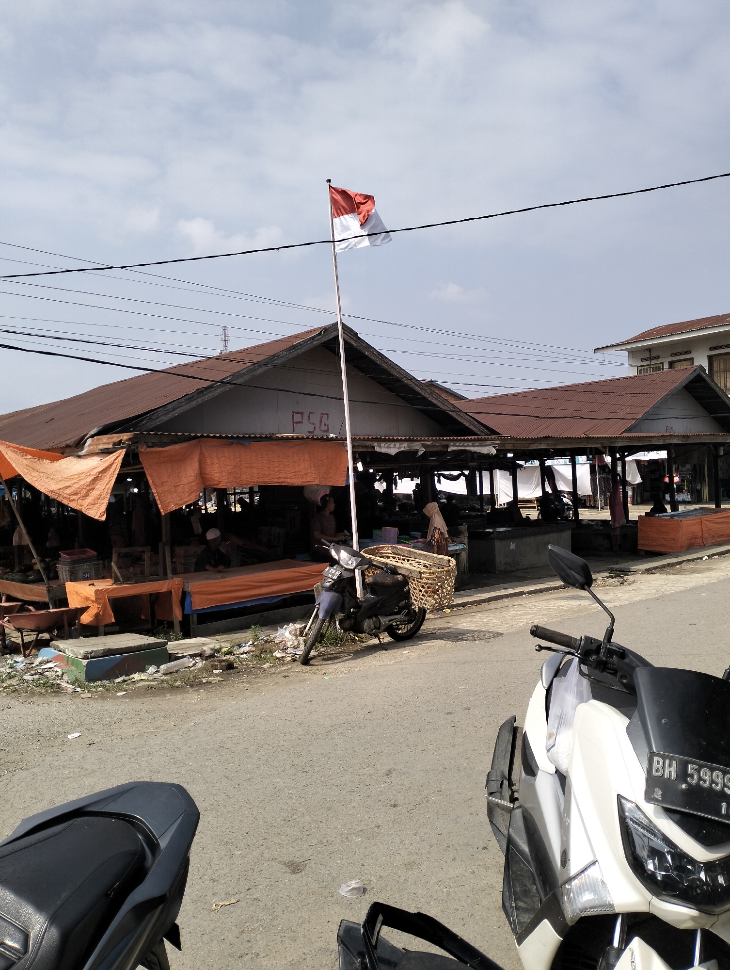 A red and white Indonesian flag flies on a tall pole in front of a covered market stall area in Serdang Jaya, Indonesia. The market stalls have corrugated metal roofs and are partially covered with orange tarps. Several people are visible within the stalls, some interacting with goods. A motorcycle with a woven basket attached is parked in front of one of the stalls. Another motorcycle is parked in the foreground, partially obscuring the view of a third motorcycle. The ground in front of the stalls is littered with debris, including plastic bottles and wrappers. Power lines are strung overhead. The sky is overcast with scattered clouds.