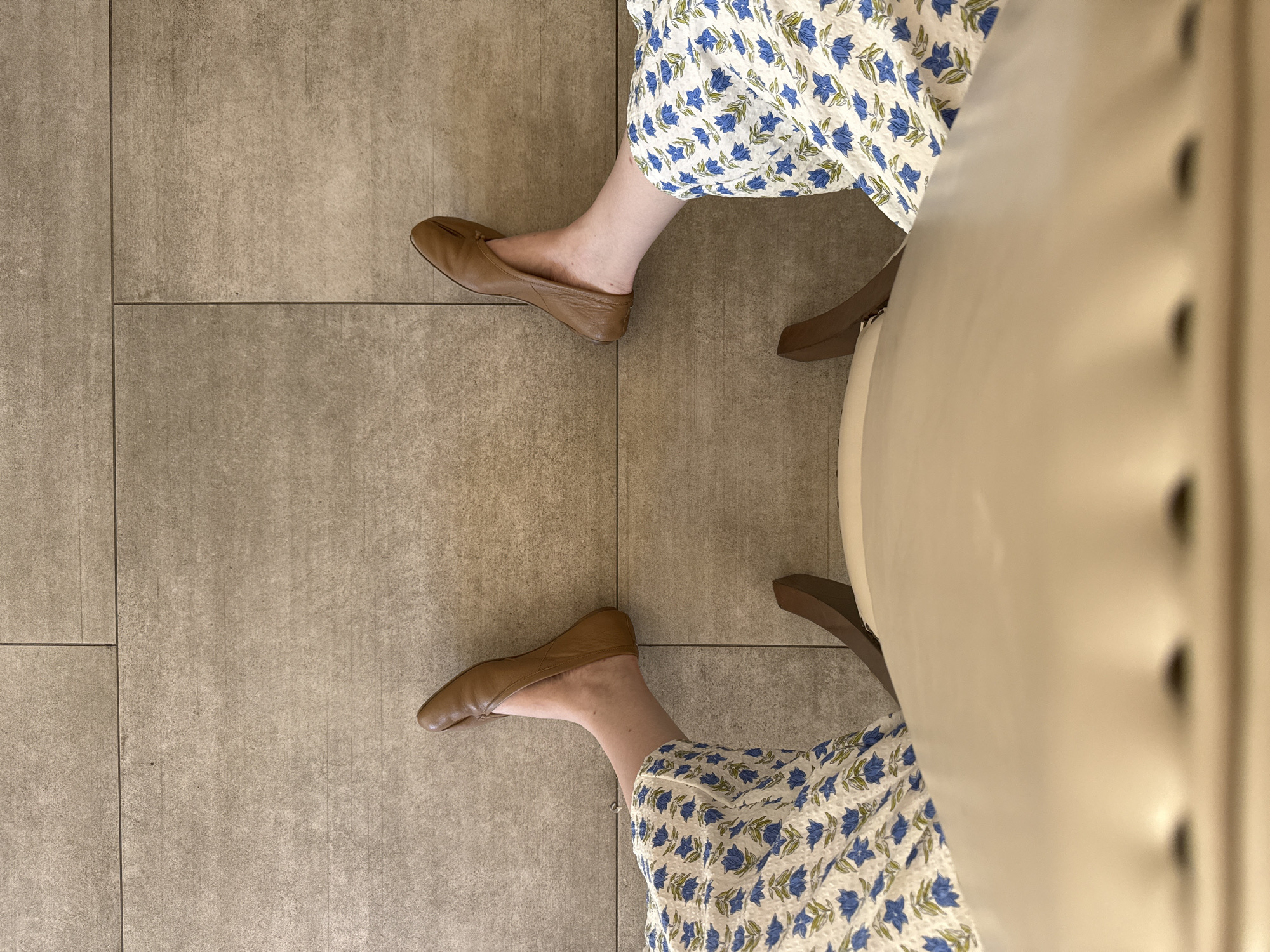The image shows a person sitting in a chair. The person is wearing a floral print dress and brown leather ballet flats. The chair is white with a tufted back. The floor is tiled. The location appears to be indoors and is unlikely to be identifiable as being in Chula Vista or any other specific location.
