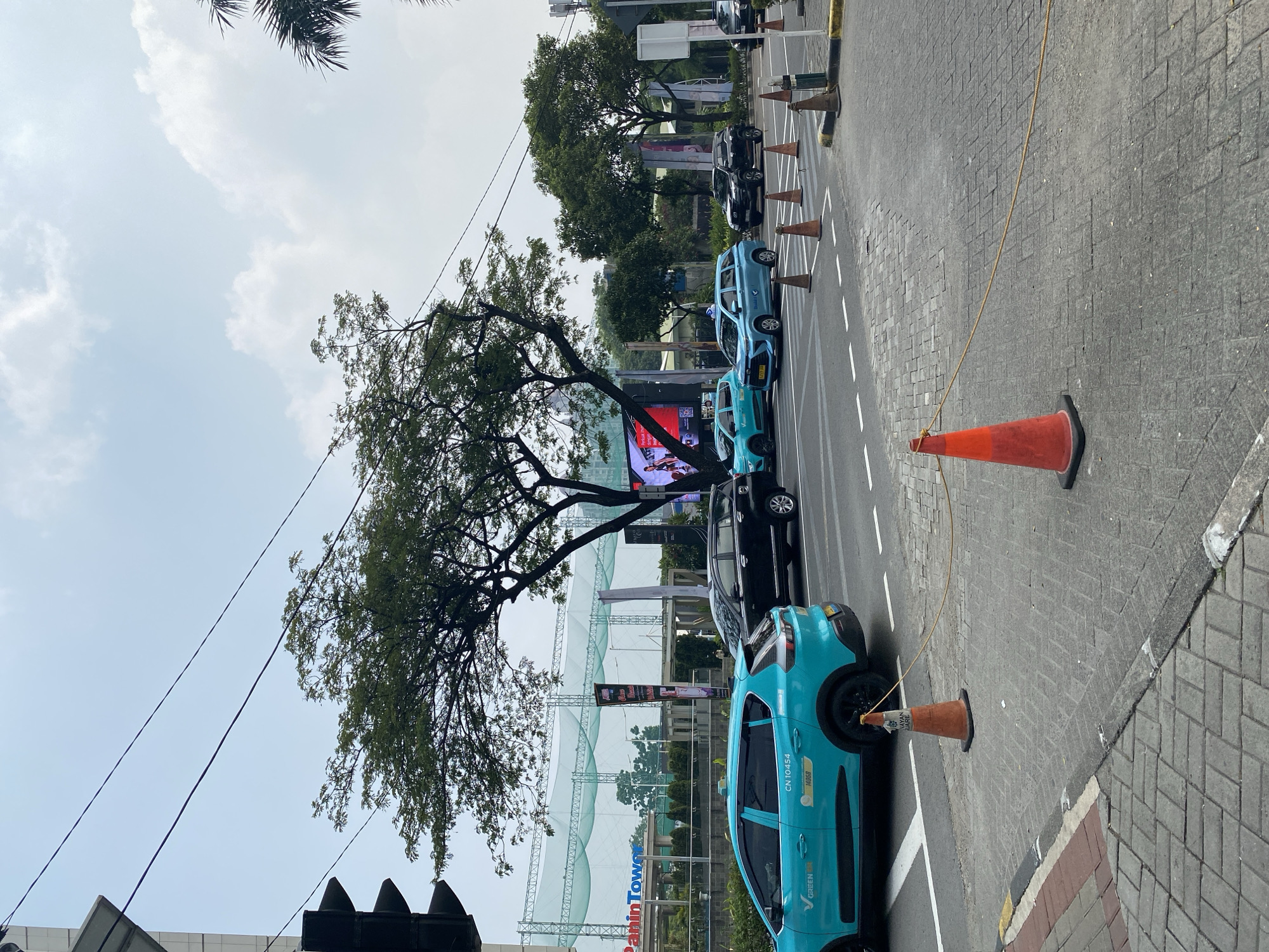The image captures a street scene in Central Jakarta, Indonesia, likely near the Panin Tower based on visible signage. Several vehicles are present on the asphalt road, including a bright teal car with "GREEN" and license plate "CN 10454" prominently displayed, suggesting it is a taxi or ride-sharing vehicle. Other cars, including a black SUV and a blue sedan, are also in motion or stopped in traffic. The road is lined with orange traffic cones connected by a rope, indicating a temporary lane closure or traffic diversion. Paved sidewalks with a brick-like pattern are visible on the left and right sides of the road. In the background, a large building with "Panin Tower" signage and a green netting structure, possibly a sports facility, are visible. Trees with dense foliage are scattered throughout the scene, and the sky is partly cloudy. A traffic light signal is mounted on a pole on the far left.