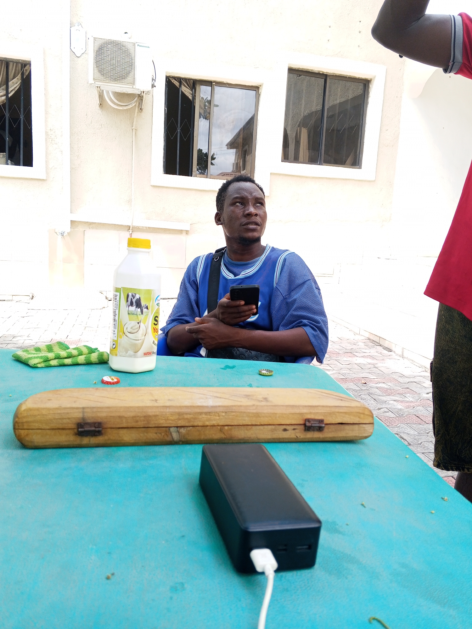 An outdoor scene features a man seated at a blue table, looking thoughtfully upwards and to his right, while holding a black smartphone. He is wearing a blue, mesh-like short-sleeved jersey and appears to have dark trousers. The partial view of another person, dressed in a red shirt, is visible on the far right of the frame.
On the teal-colored table, several items are present. A tall, white plastic bottle with a yellow cap, labeled "SO YOGURT" and "NAFDAC Reg.", stands near the man, featuring an illustration of a cow and a glass of yogurt. Adjacent to it are a green and white striped cloth or towel and a red bottle cap. In the foreground, a rectangular wooden box with metal hinges and clasps rests on the table, alongside a black power bank with a white charging cable connected to it.
The background reveals a light-colored building, possibly white or cream, with two distinct windows. Both windows are fitted with dark security grilles and have reflective glass. An outdoor air conditioning unit, bearing the "Panasonic" brand name, is mounted on the building wall between the two windows. The ground beyond the table is paved with patterned tiles.
The setting appears to be a casual outdoor area, possibly a patio or courtyard, during bright daylight hours, as suggested by the clear lighting and shadows. The man's gaze implies he is observing something or someone beyond the camera's view, creating an impression of a momentary pause or interaction in a relaxed environment.