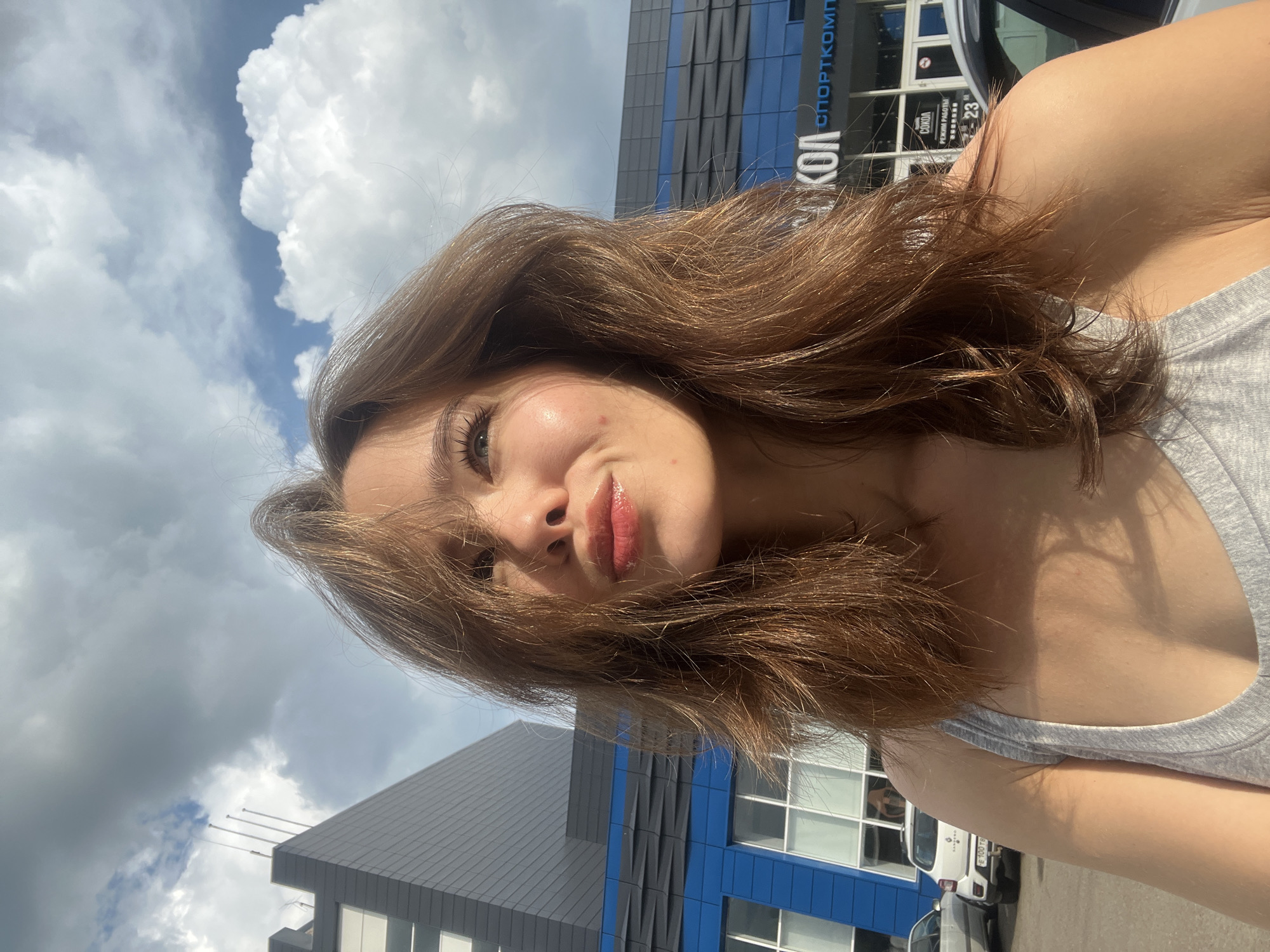 The image shows a young woman with shoulder-length brown hair posing for a selfie outdoors. She is wearing a grey tank top. Her head is tilted upwards, looking towards the sky, and her lips are slightly puckered. Behind her, a modern building with blue and grey accents is visible, with the Cyrillic text "СОКОЛ СПОРТКОМПЛЕКС" (Sokol Sports Complex) displayed prominently on its facade. The building appears to be a sports facility. Cars are parked in front of the building. The sky is partly cloudy with bright sunlight casting shadows. This location is identified as the Sokol Sports Complex in the city of Krasnoyarsk, Russia.