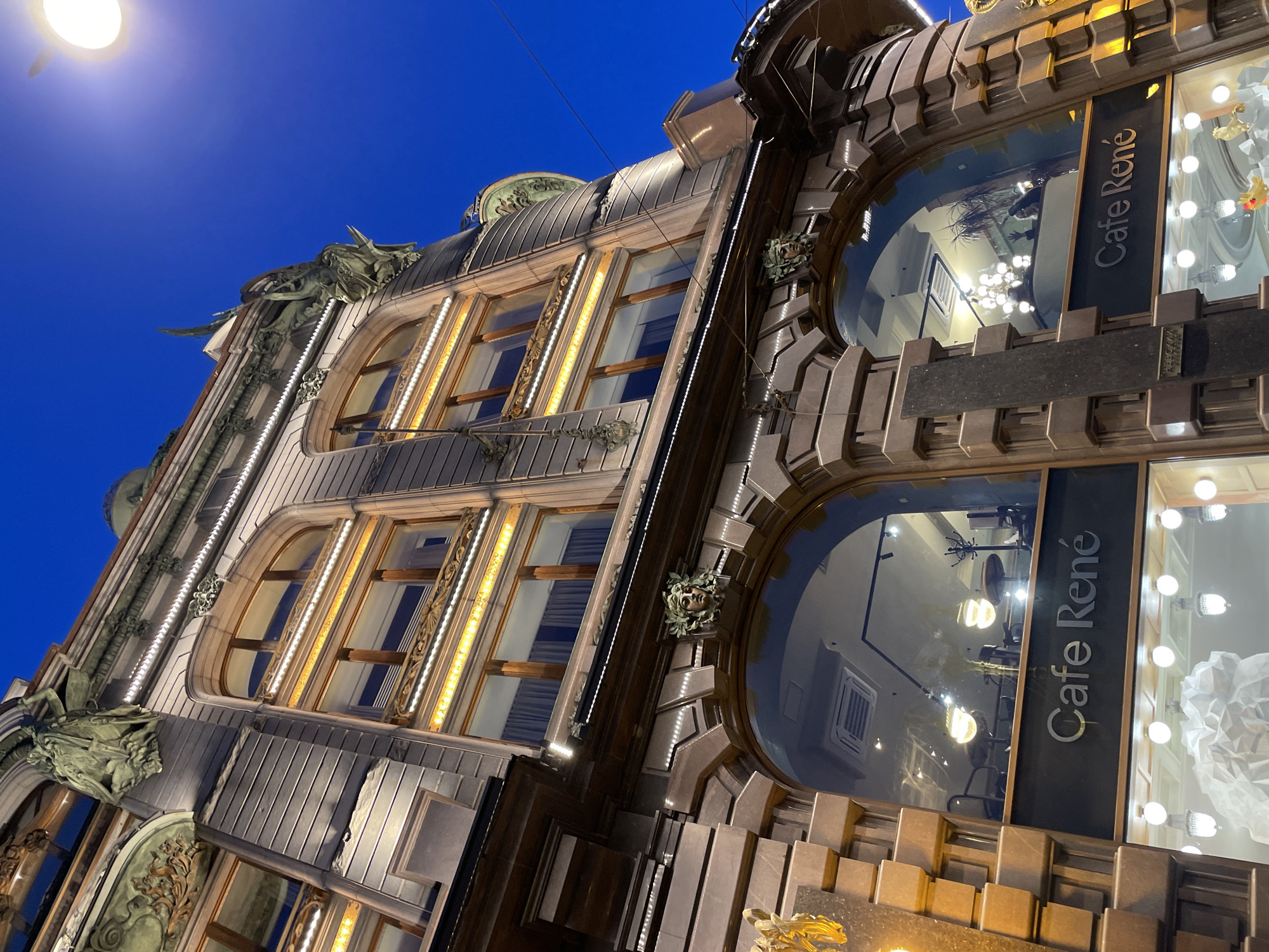 An upward-angled shot captures the illuminated facade of an ornate building at night, set against a deep indigo sky. The architectural style appears to blend elements of Art Nouveau and early 20th-century design, characterized by intricate details, sculptural elements, and decorative window frames.
The building is brightly lit by a combination of interior lights and external illumination. Vertical strips of warm, yellow-orange LED lights accentuate the architectural lines on the left side of the facade, highlighting several windows with arched tops and elaborate surrounds. Below these windows, dark, sculptural forms, possibly depicting mythological figures or gargoyles, are integrated into the stonework. A bright streetlight is visible in the upper left, emitting a glow against the dark sky.
On the right side of the image, two large, curved display windows reveal the interior of a establishment. The name "Cafe René" is prominently displayed in white, sans-serif font on a vertical dark panel between these windows. Inside, numerous spherical light fixtures emit a bright glow, and reflections on the glass allow glimpses of a contemporary interior with tables and chairs. The facade around these display windows features geometric, stepped patterns and smaller decorative elements, some resembling sculpted faces or masks adorned with foliage. Overhead wires are faintly visible crossing the sky. The overall impression is one of a grand, historically significant building beautifully preserved and lit, located in an urban setting like Saint Petersburg, Russia, as suggested by the context.