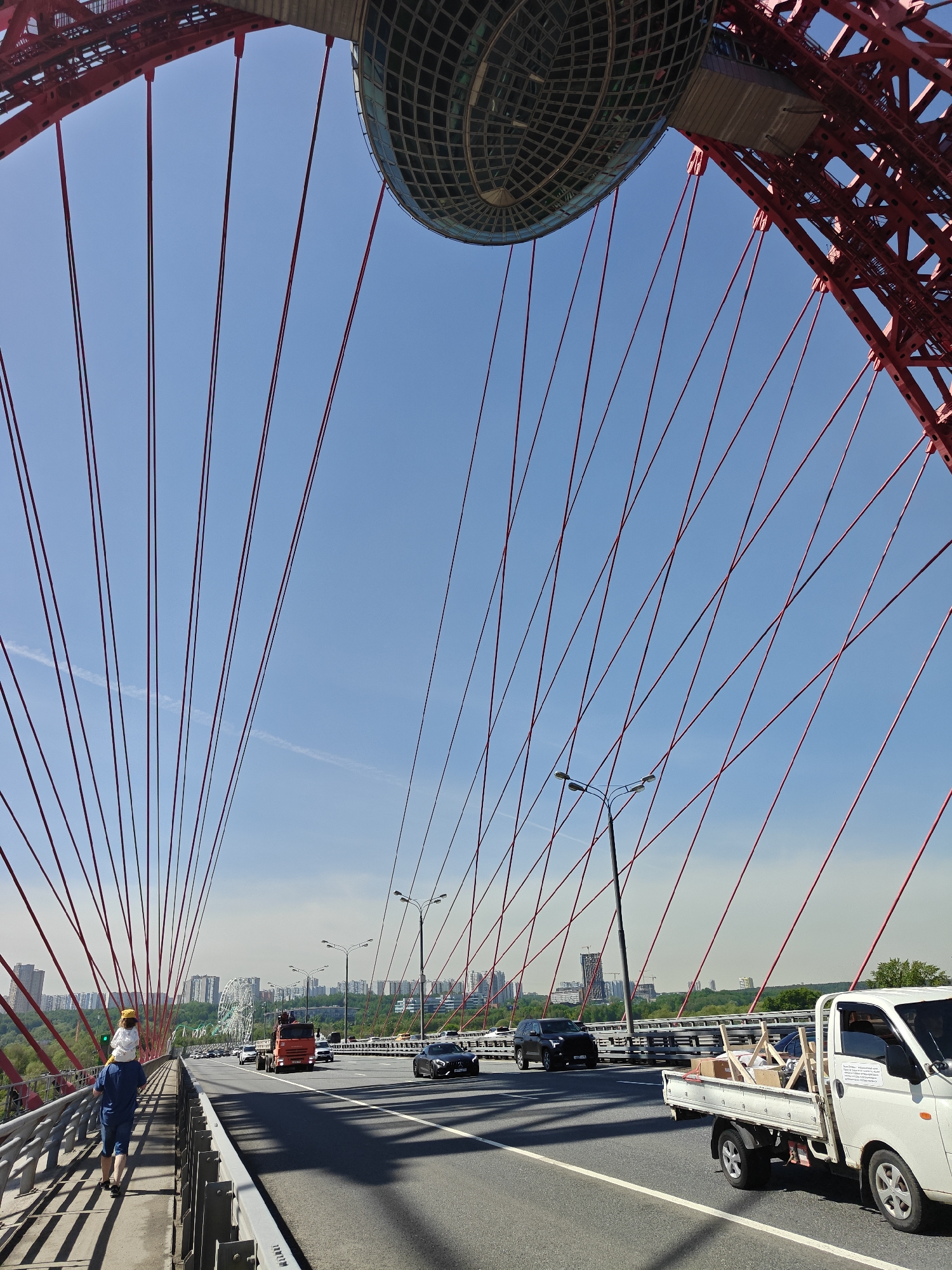 A daytime, low-angle shot shows a pedestrian bridge in Khimki, Russia, with red support cables stretching towards a central, round observation deck. A man carries a child on his shoulders, walking along the pedestrian walkway on the left. Cars and trucks travel on the adjacent roadway. Street lamps line the road. In the background, buildings are visible against a clear blue sky.