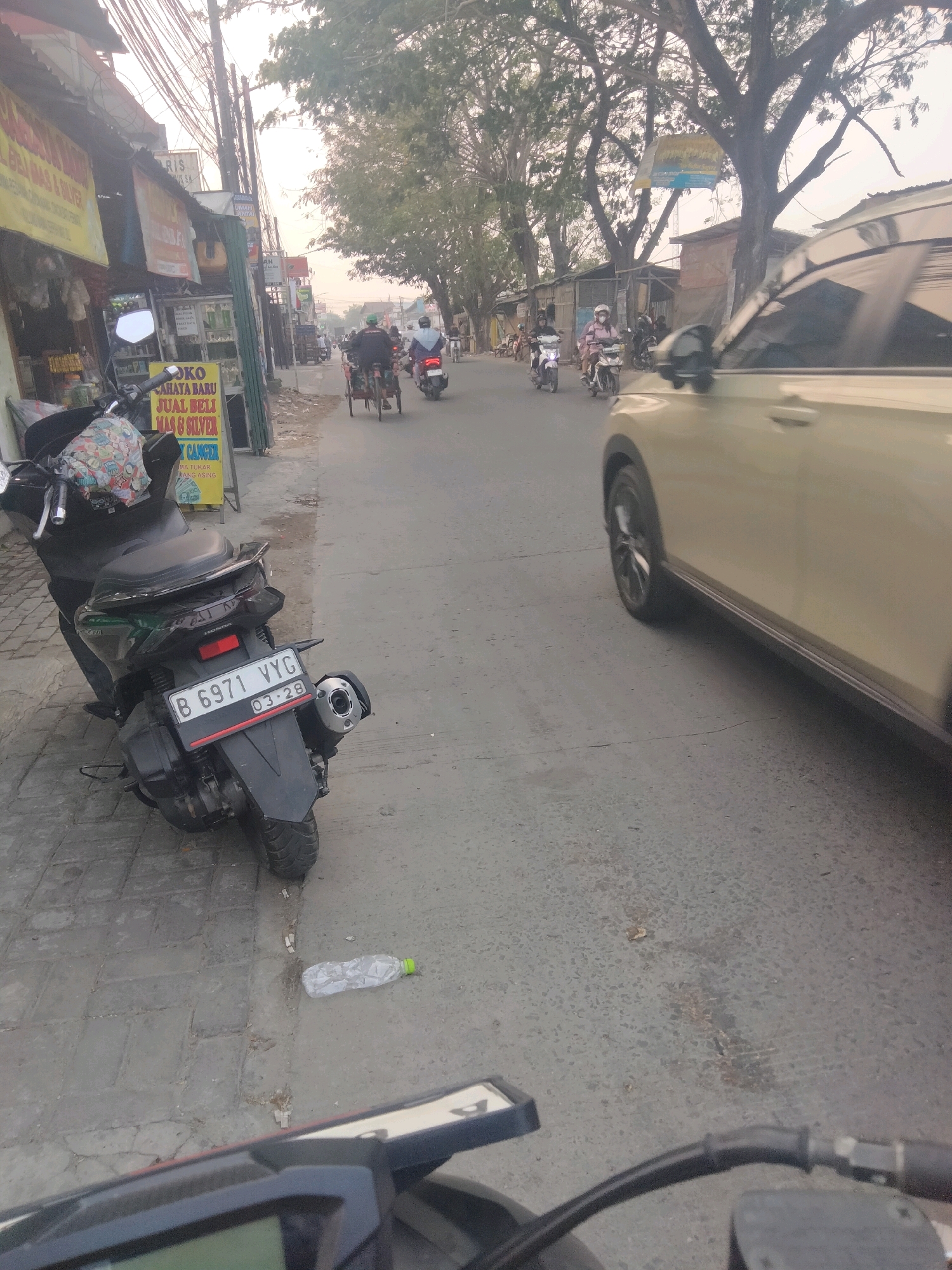 A scene is captured on a street in Sepatan, Indonesia, likely during the day given the lighting conditions. The foreground is dominated by the rear of a black motorcycle, which is parked on a paved sidewalk. The motorcycle has a license plate that reads "B 6971 VYG" and shows the expiry date "03-28". A piece of fabric with a colorful pattern is draped over the motorcycle's seat.
To the left of the motorcycle, a business establishment is visible. A prominent yellow sign displays text that appears to be a price list or advertisement, mentioning "JUBL BELI MAS & SILVER" and "CANCER". Various other signs and posters are affixed to the building.
The street itself is an asphalt road with some visible cracks and debris, including a discarded plastic water bottle with a green cap lying on the sidewalk. Traffic is present on the road. Several motorcycles, a bicycle rickshaw, and a light-colored car are depicted. The car is positioned on the right side of the frame, partially obscuring the view of the road and sidewalk. On the motorcycles, riders are visible, some wearing helmets. One rider on a rickshaw is also present.
The environment suggests a typical urban or suburban street with trees lining the sides of the road, their branches reaching over the asphalt. Utility poles with a dense network of wires are also prominent. The overall atmosphere is that of a busy street with ongoing activity.