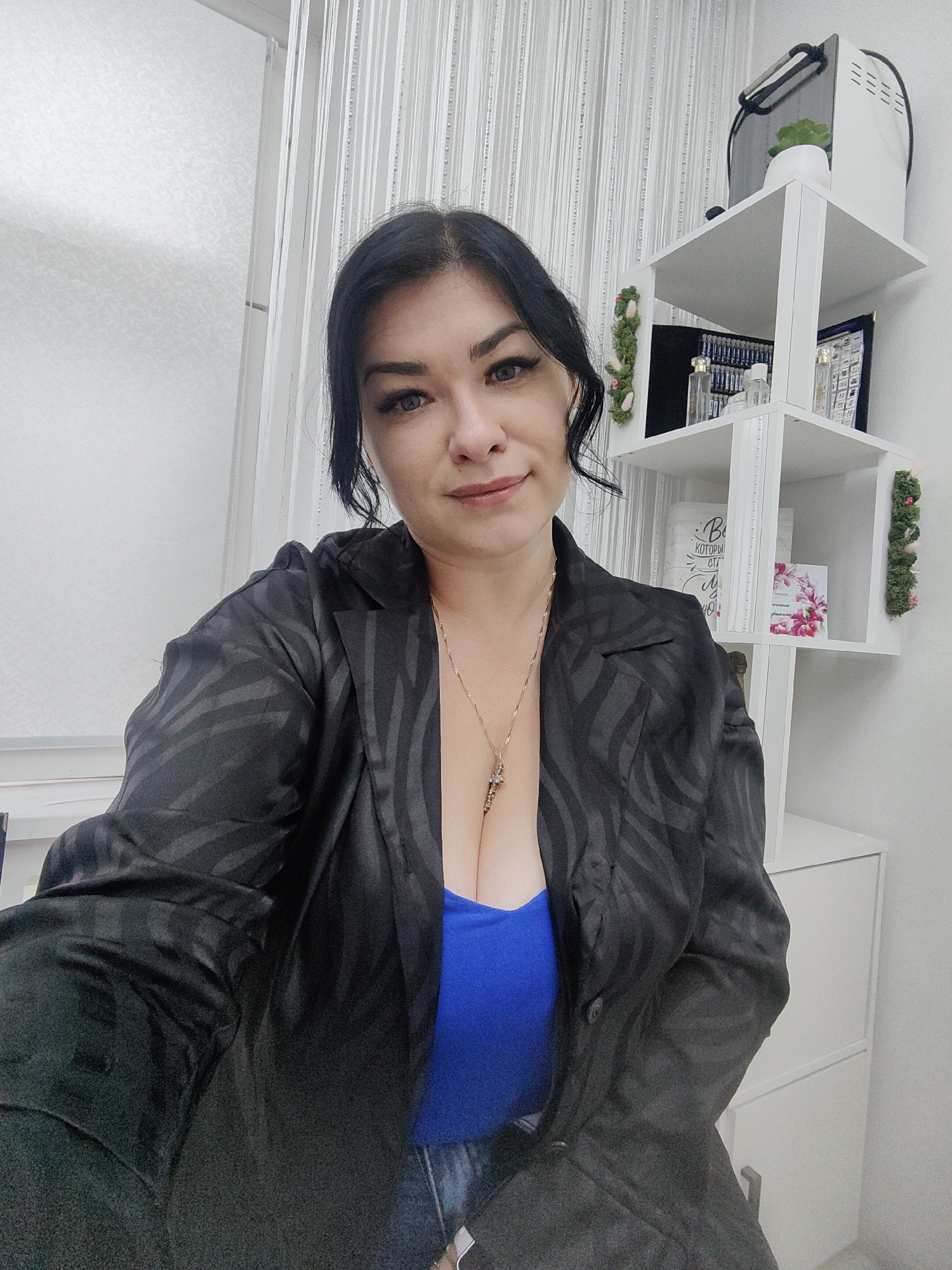 A medium shot depicts a woman with dark hair wearing a black blazer over a blue top and jeans. A gold necklace hangs around her neck. The background includes a white shelf unit with various items, including a small plant and decorative moss, as well as a window with light-colored curtains. The setting may be indoors, possibly in a business or home environment. There is no explicit indication of the city of Nizhnevartovsk, Russia.