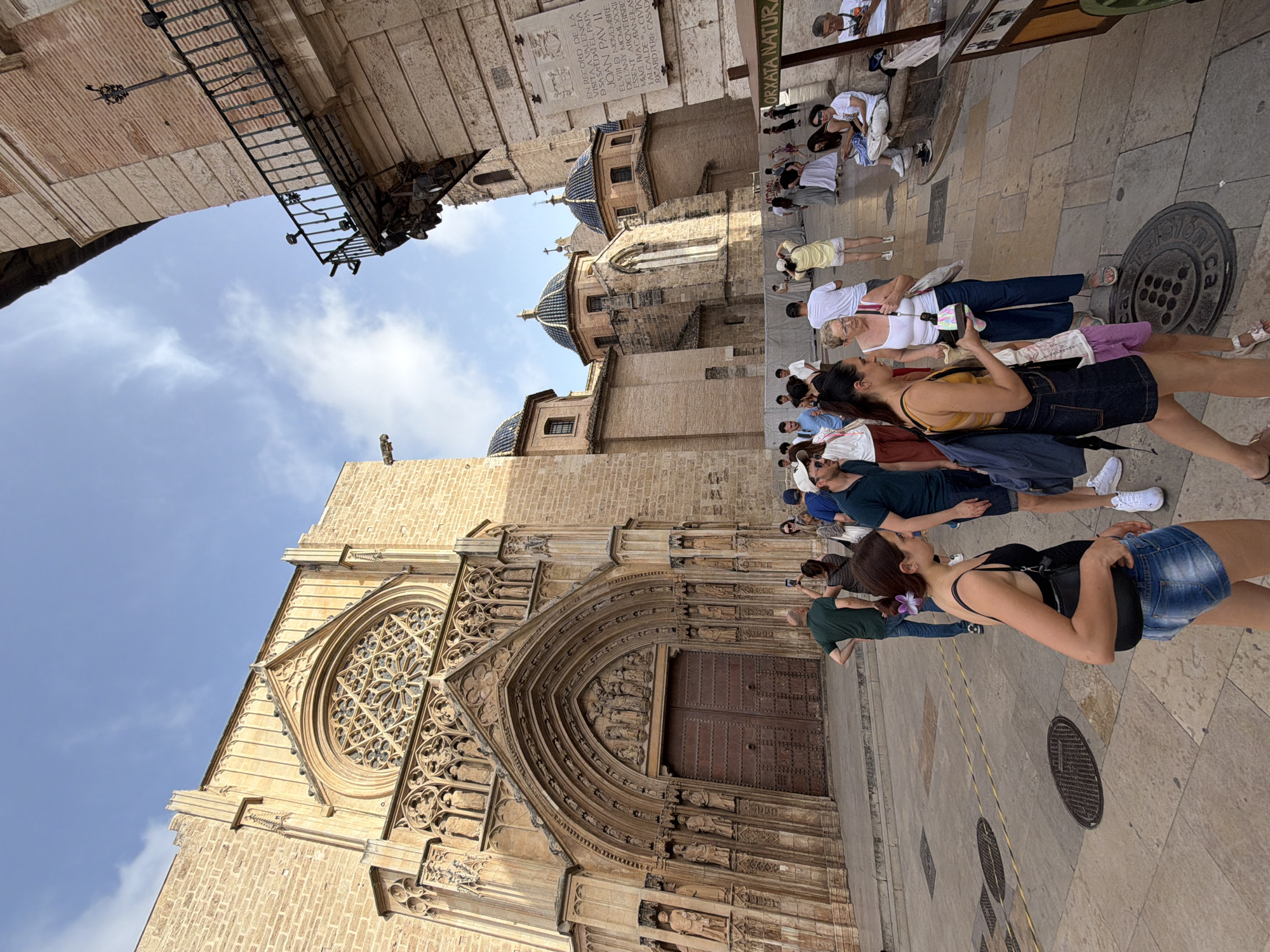 In an outdoor plaza, a large, ornate stone cathedral with a circular stained-glass window above its arched entrance dominates the left side of the frame. The facade is intricately carved with statues and architectural details. To the right of the cathedral, a lighter-colored brick building with a wrought-iron balcony juts into the frame, casting a shadow. The ground is paved with cobblestones and concrete, with manhole covers visible. A crowd of tourists and locals are dispersed across the plaza. Several individuals are walking, some are taking pictures, and others are conversing. In the foreground, a young woman in denim shorts and a black tank top walks towards the right, holding a black purse, with a purple flower pinned in her hair. Behind her, an older woman in a white tank top and blue pants speaks to her. Further back, a man in a green shirt is taking a photo with his phone, and a group of people in casual attire are visible near the cathedral entrance and the brick building. The sky above is partly cloudy with patches of blue. A street vendor's cart is partially visible on the far right. The scene captures a moment of tourism and daily life in an urban historical setting.