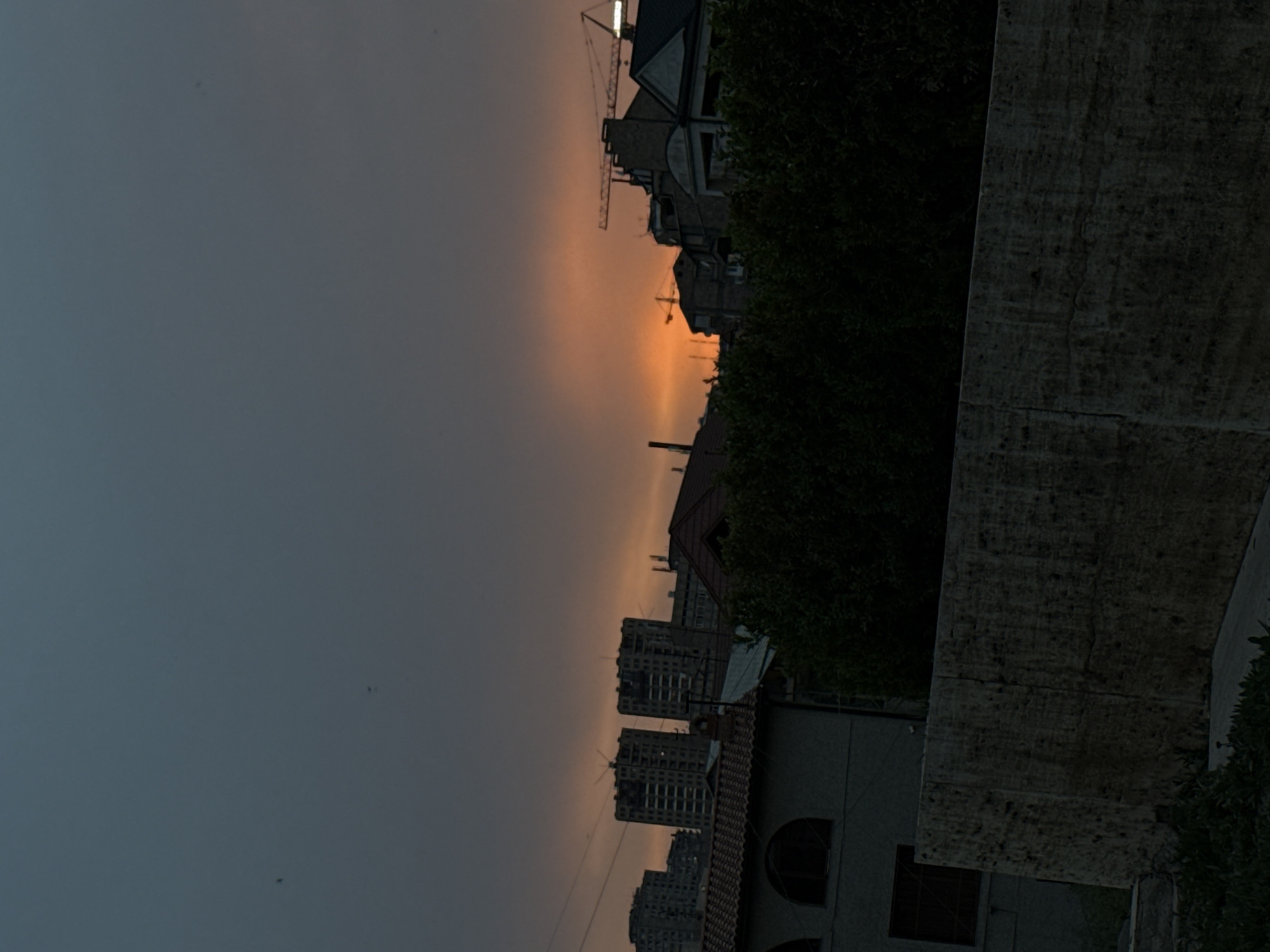 The image shows a cityscape at dusk, likely in Yerevan, Armenia, given the architecture and setting sun. The sky transitions from dark gray in the lower left to a soft orange horizon, indicating the sunset. Buildings of varying heights are visible, including taller apartment complexes and smaller residential structures. Construction cranes dot the skyline, suggesting ongoing urban development. Vegetation is present, adding a touch of green to the scene. A wall is in the foreground.