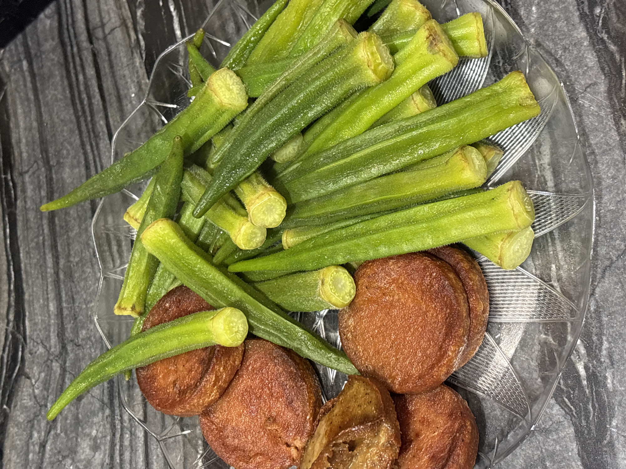 A clear glass plate containing fried okra and sausage patties sits on a dark gray marble countertop. The okra pods are bright green, with fuzzy exteriors and cut ends revealing a lighter green interior. The sausage patties are round and a deep reddish-brown, with some appearing slightly charred. The countertop has a textured, marbled pattern. The scene is illuminated from above, casting shadows. There are no people or other objects visible in the image. The location is not specified to be in Dubai, United Arab Emirates.