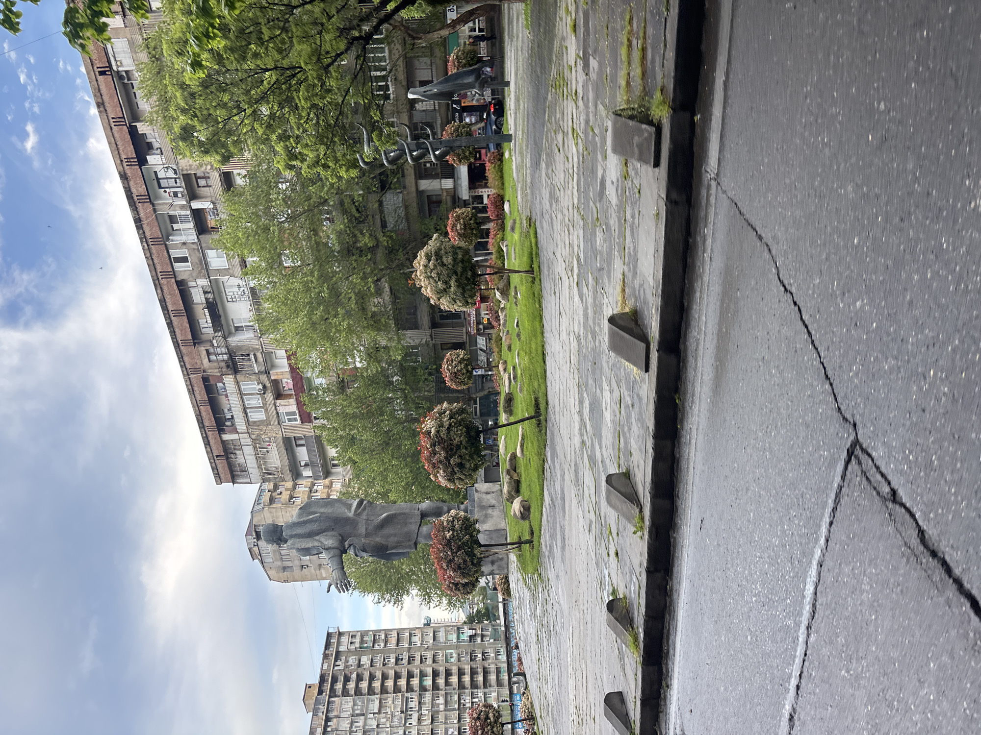 The image shows a view of the Melikishvili Avenue in Tbilisi, Georgia. It is taken at street level, looking towards a park area with a statue and trees. The statue appears to be of a man, standing in a public square. In the background, there are residential buildings. The street is made of asphalt, and there is a sidewalk made of stone slabs adjacent to the street. The sky is partially cloudy.