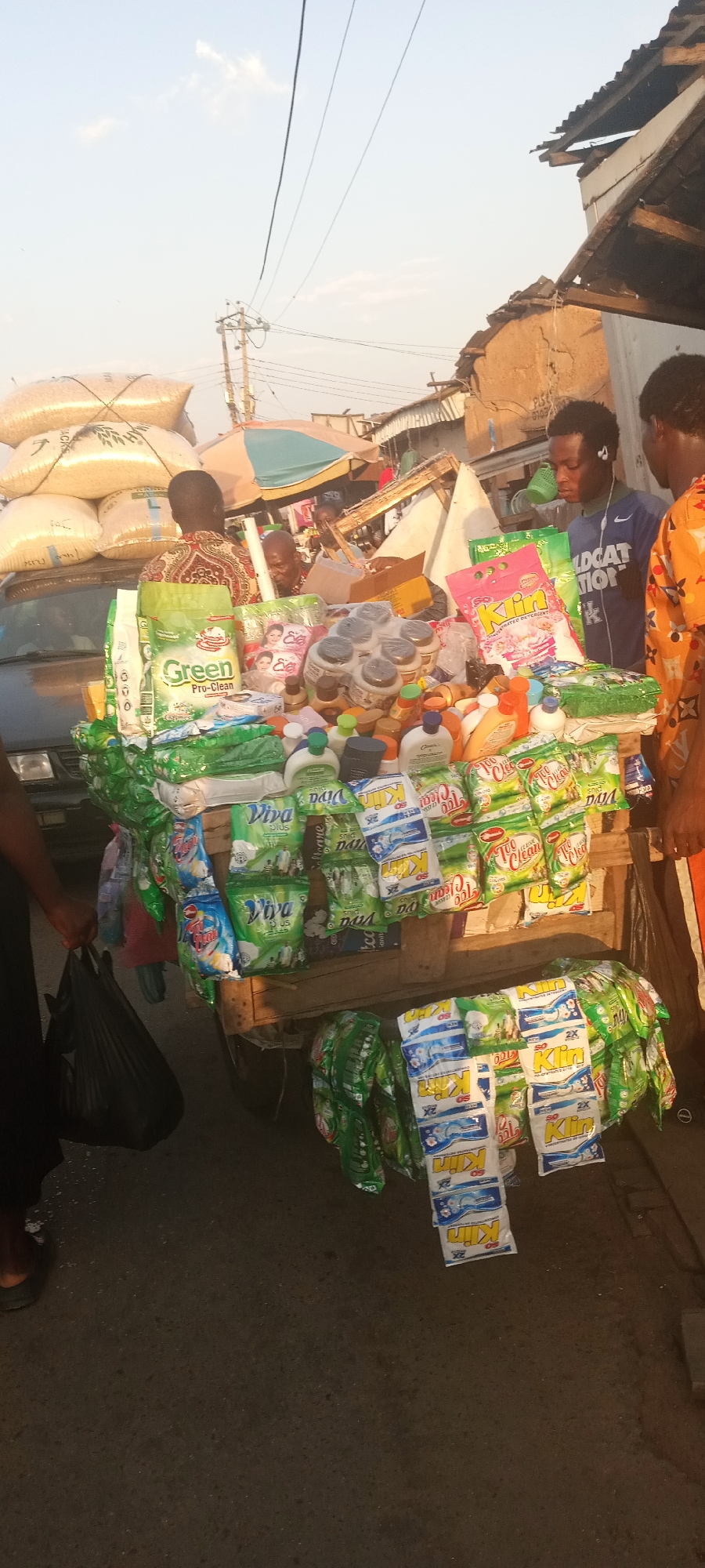 Here is a description of the image:
A street vendor in Jos, Nigeria, displays a variety of cleaning products on a wooden cart. The cart is laden with stacks of detergent powders (brands like "Klin," "Too Clean," and "Viva Plus"), dish soaps, and what appear to be skin lotions or creams (brand "Eva"). Some of the products hang from the cart. Bags of rice are loaded on a vehicle behind the seller's cart. A customer wearing headphones looks at the merchandise with another man in an orange shirt. Another person holds a black shopping bag, seemingly in the process of purchasing items. The scene appears to be a busy market or street. In the background are other shops, stalls, and people. The lighting suggests it is either early morning or late afternoon.