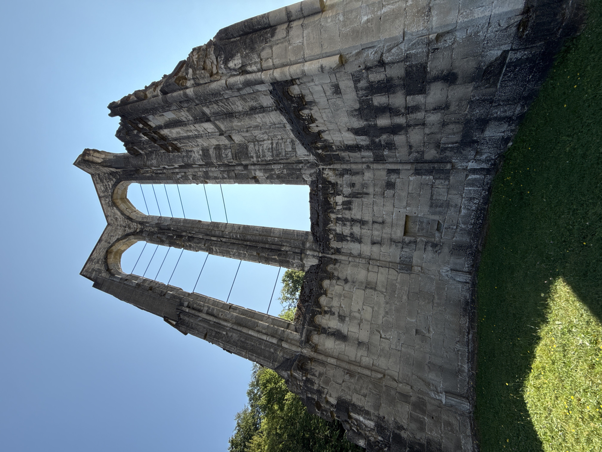 The image shows a low-angle view of the ruins of the Walkenried Abbey in Walkenried, Germany. The ruins are made of weathered stone blocks and feature large arched openings against a clear blue sky. A lush green lawn with scattered yellow wildflowers is in the foreground, with the shadows of the ruins cast upon it. Trees can be seen on the left side of the image, bordering the ruins. The structure appears ancient and dilapidated, with missing sections and visible wear on the stonework.